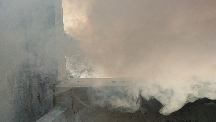The image displays the interior of a concrete structure, presumably located in Dhaka, Bangladesh, heavily permeated by dense smoke. The left side of the frame features a grey concrete wall exhibiting several linear cracks, with a horizontal concrete beam spanning across the upper left. A vertical concrete structural element forms a corner, opening to a heavily smoke-filled area on the right. This area is obscured by a large volume of light grey to brownish-orange smoke, within which scattered faint bright points are discernible, potentially indicating embers or reflections of an external light source. The air inside the visible portion of the structure is actively filled with this smoke. No individuals or identifiable specific objects beyond the concrete architecture and smoke are visible.