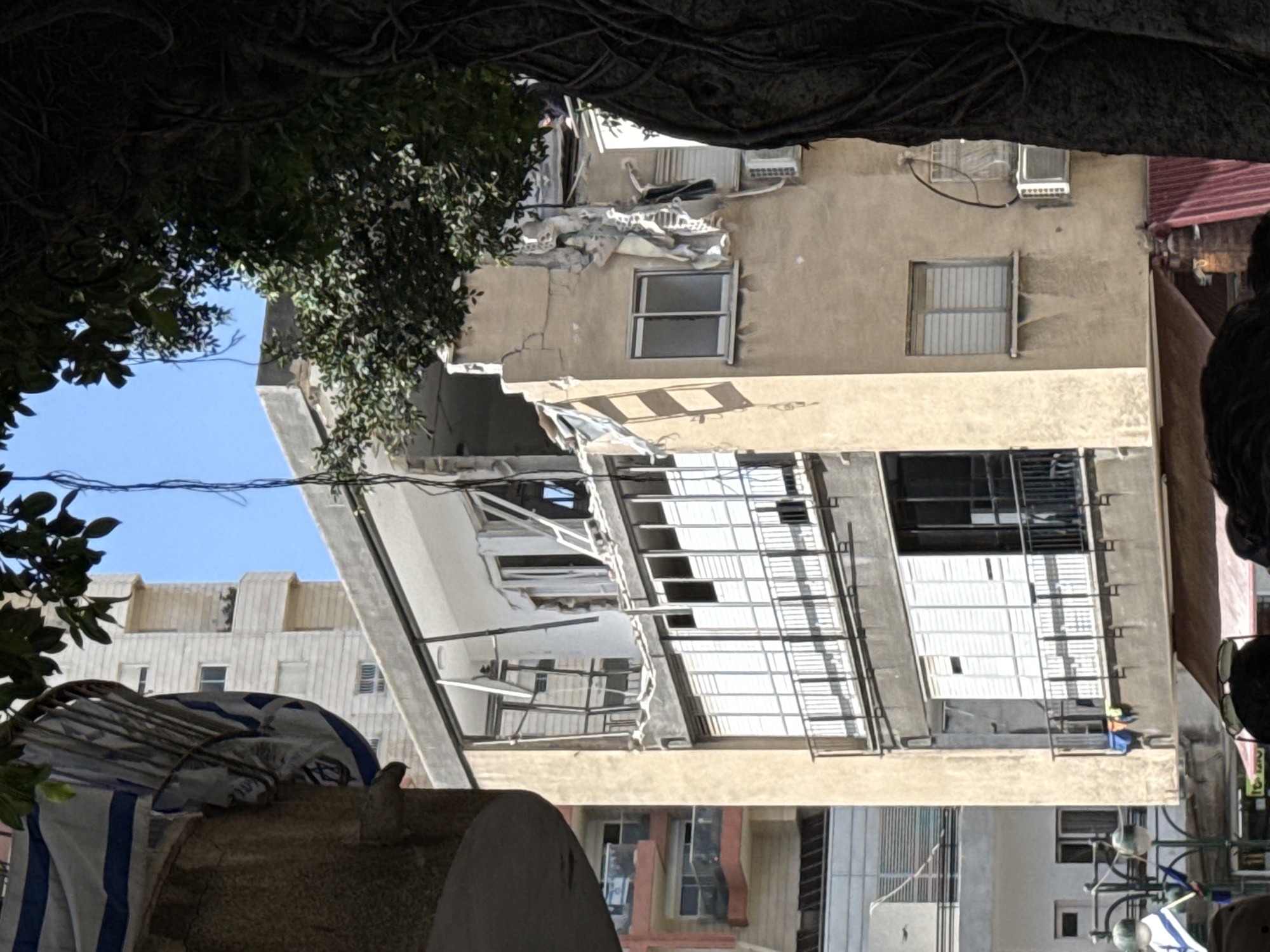 In Ramat Gan, Israel, a severely damaged building shows extensive structural failure. A section of the building's facade has collapsed, revealing the interior framework. Debris is scattered around the damaged area. Several windows on the building are visible, with some appearing intact while others are broken or missing. The building's exterior is weathered and shows signs of age. The image is framed by foliage from a tree. The building is surrounded by other structures. The environment appears urban, with signs of human activity in the vicinity.