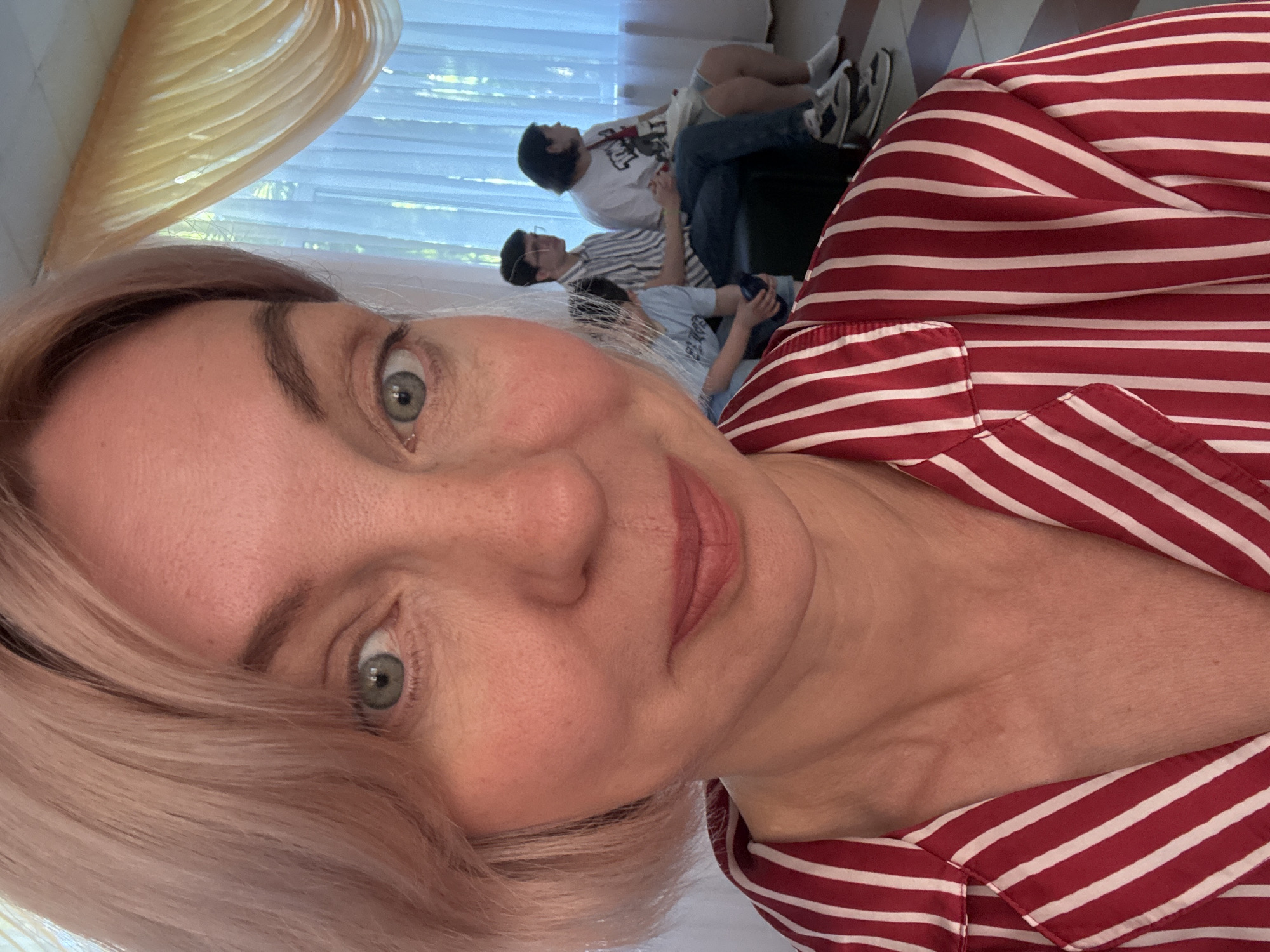 A woman with light blonde hair, light blue-green eyes, and a gentle smile is captured in a close-up selfie, looking directly at the camera. She wears a red and white striped shirt or pajama top, which is a prominent feature in the foreground. Her face, showing a fair complexion, fills the left side of the frame, with some hair strands framing her features.
In the blurred background, three younger individuals are seen in an indoor setting, appearing relaxed. One person in a light blue t-shirt seems to be holding an object in their hands. Another wears a white and blue striped shirt and glasses, looking downwards. The third individual, in a white t-shirt, jeans, and sneakers, is seated on a dark couch or chair, also appearing to be engaged with something in their hands.
The scene is illuminated by bright natural daylight coming through a window with light-colored curtains or blinds, indicating it is daytime. The overall mood is casual and comfortable. No visible text is present in the image. The location context provided is Sochi, Russia.