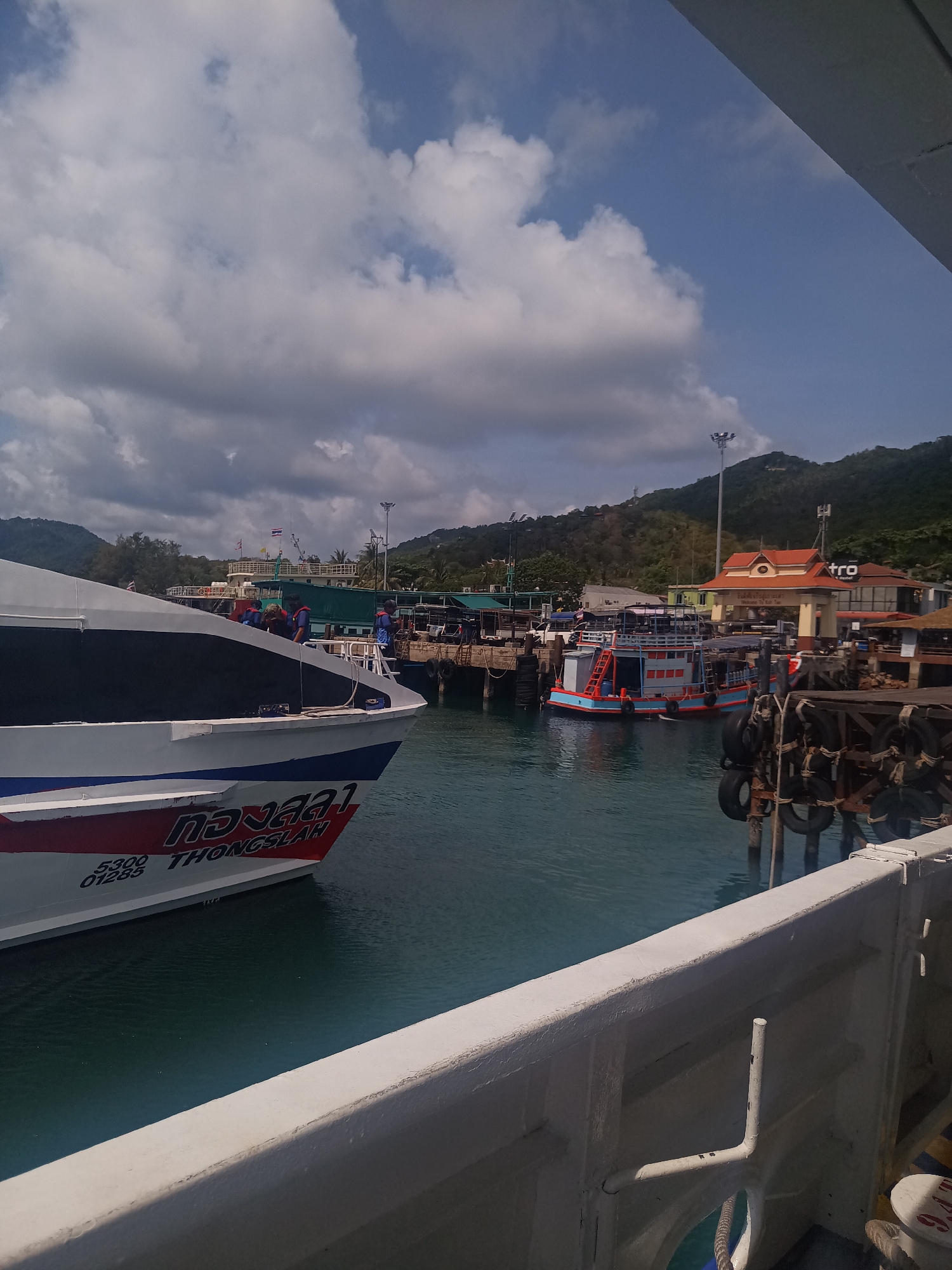 The image depicts a harbor scene in Tambon Ko Tao, Thailand, on a partly cloudy day. The foreground features a white boat with "Thongsala" written on its bow. Adjacent to it is the white railing of another boat, from which the photo appears to have been taken. In the middle ground, there's a wooden pier with several boats docked, including a green ferry and a smaller orange and blue passenger boat. On the pier, there are structures of varying sizes and purposes, including a building with a red roof and the "Metro" sign. The background includes a lush green hillside and the blue sky with grey clouds.