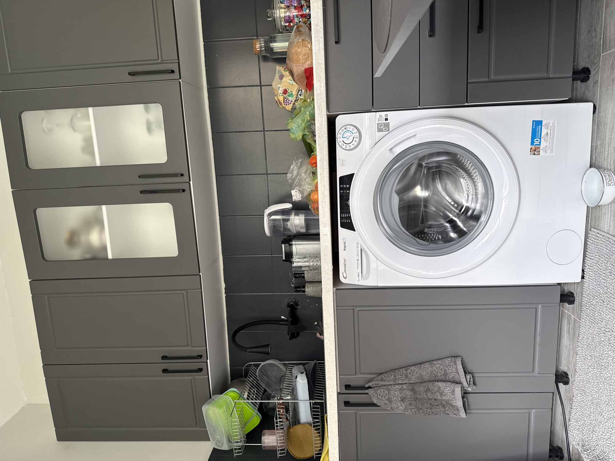 The image shows a kitchen interior. The primary elements are two sets of grey kitchen cabinets, one with frosted glass panels and the other without. Between the cabinets, there is a narrow space containing food items, a vacuum cleaner, a kitchen faucet, and a dish rack. Adjacent to the cabinets on the right is a white washing machine. The location is likely a private residence within Stara Zagora, Bulgaria, based on the language of the washing machine label.