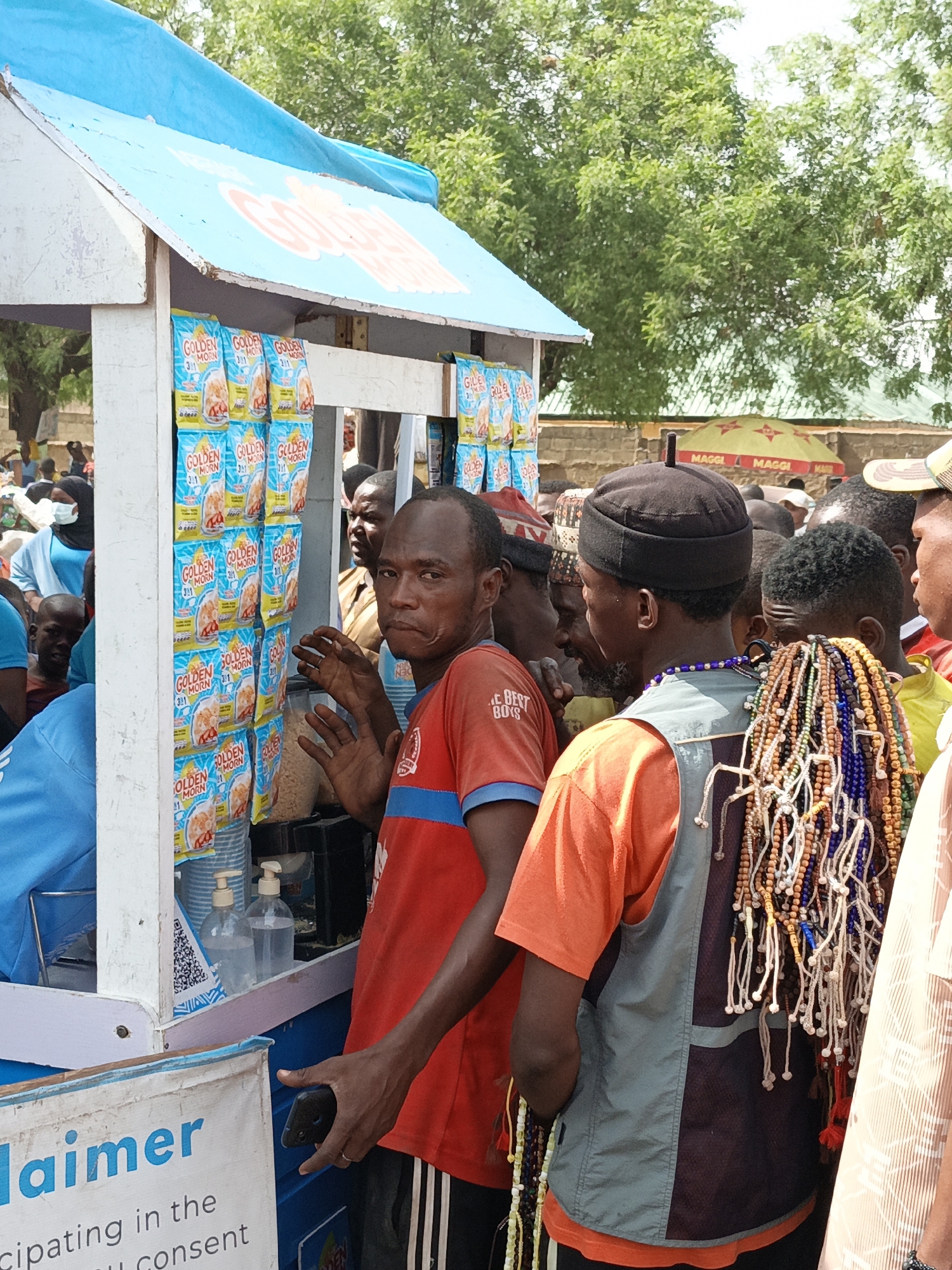 A medium shot captures a food vending stand in Kawaji, Nigeria, surrounded by a crowd. The vending stand, under a blue awning displaying "GOLDEN MORN," has packages of "GOLDEN MORN" cereal prominently displayed. A man in a red shirt with black-and-white striped pants gestures towards the stand with one hand and holds a phone in the other. Another man, wearing an orange shirt and a grey vest, has strands of colorful beads hanging on his back. The crowd behind them appears diverse, with people in various traditional and casual clothing. In the background, an umbrella with "MAGGI" branding is visible. The environment appears to be an outdoor public space.