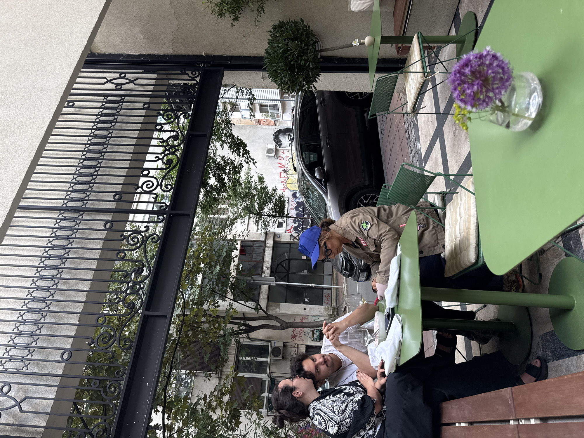 The image captures an outdoor scene, likely at a cafe in Belgrade, Serbia. Three individuals are present: two are seated closely together, while the third, wearing a blue cap and a brown jacket, is seated across from them. They appear to be looking at something on a table, possibly a phone. The table is green, and a vase with purple flowers sits on it. The background features a street with cars parked and buildings with graffiti. A decorative black metal gate is visible on the left side of the frame, and green tables and chairs are lined up along the pavement in front of the building. The time of day appears to be daylight.
