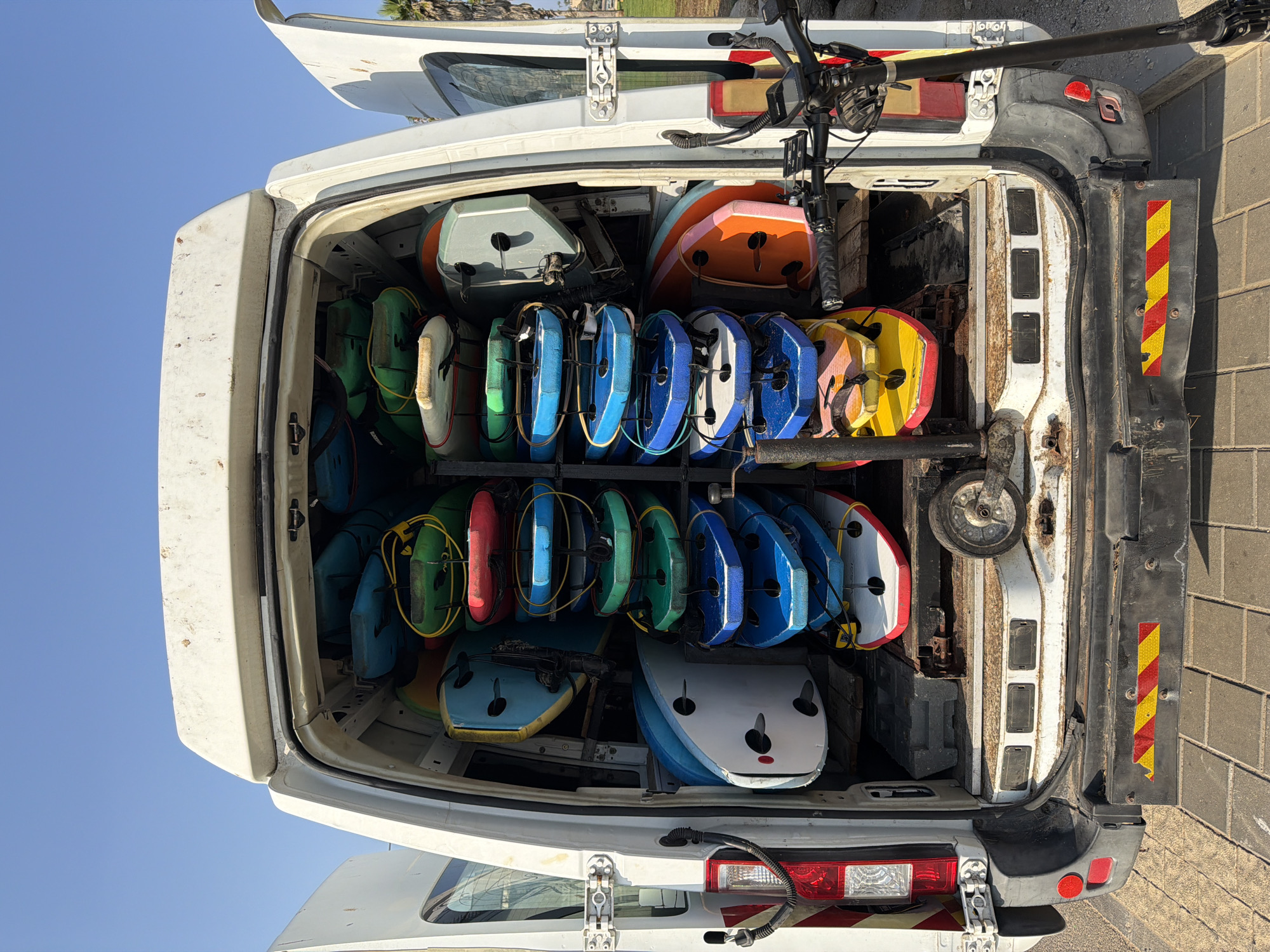 A white van with its rear doors open is parked on a paved area outdoors, likely in Tel-Aviv, Israel, indicated by the clear blue sky and palm trees visible in the background. The van is filled with numerous brightly colored surfboards and bodyboards, neatly stacked on racks. These boards are in various shades of blue, green, red, yellow, orange, and white, with some featuring contrasting accents. A black electric scooter with a display screen is parked to the right of the van, partially obscuring the van's rear bumper. The van's bumper has reflective red and yellow stripes. The environment appears to be sunny and warm.