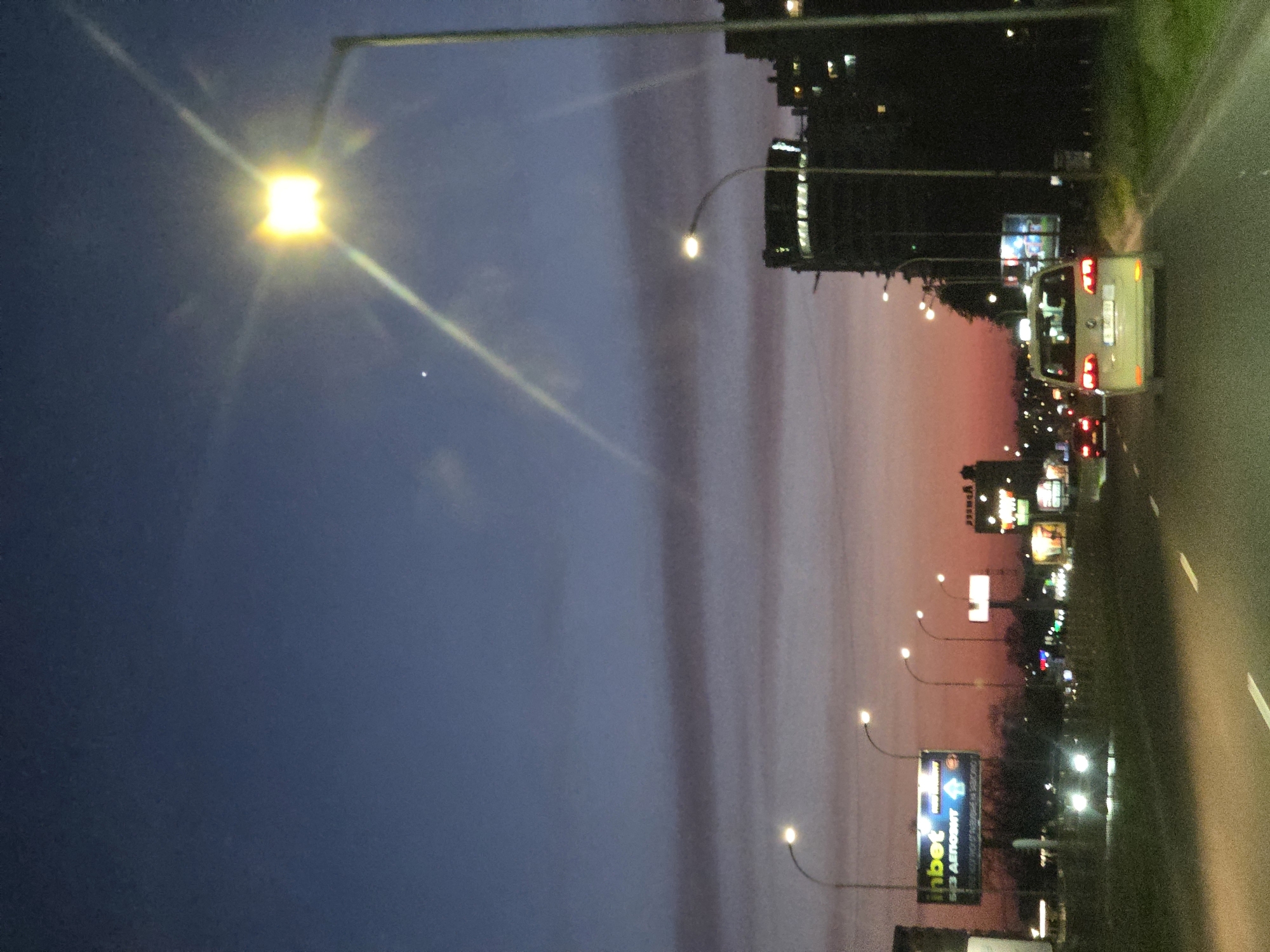 The image shows a nighttime scene along a road in Varna, Bulgaria. The sky transitions from dark blue to shades of pink and orange near the horizon. A tall street lamp is visible on the left side, emitting a bright light. A road runs along the right side of the image, with a white lane marker. On the right side of the road, there are multiple light sources, including street lamps and illuminated billboards, as well as the lights of buildings. A white van and the rear lights of other vehicles are visible on the road. One of the billboards is labeled "Inbet". The presence of buildings and billboards suggests an urban or suburban setting.