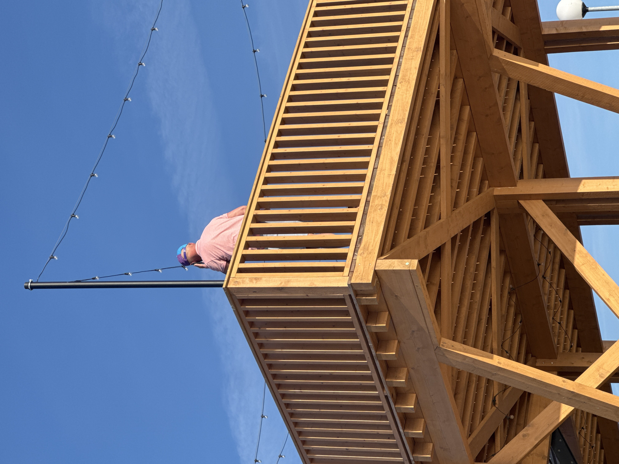 An over-the-shoulder shot captures a person wearing a pink collared shirt and a blue and purple baseball cap, standing on a wooden observation deck, looking towards the sky. They appear to be holding a phone to their ear, possibly engaged in a conversation. The deck features a wooden railing made of vertical slats. The background is a clear blue sky with a few wispy clouds. A black pole with a string of lights runs diagonally behind the person. The underside of the deck reveals a complex wooden support structure. The overall scene suggests an outdoor setting, potentially a public area within the city of Tallinn, Estonia, like a promenade or a viewpoint.