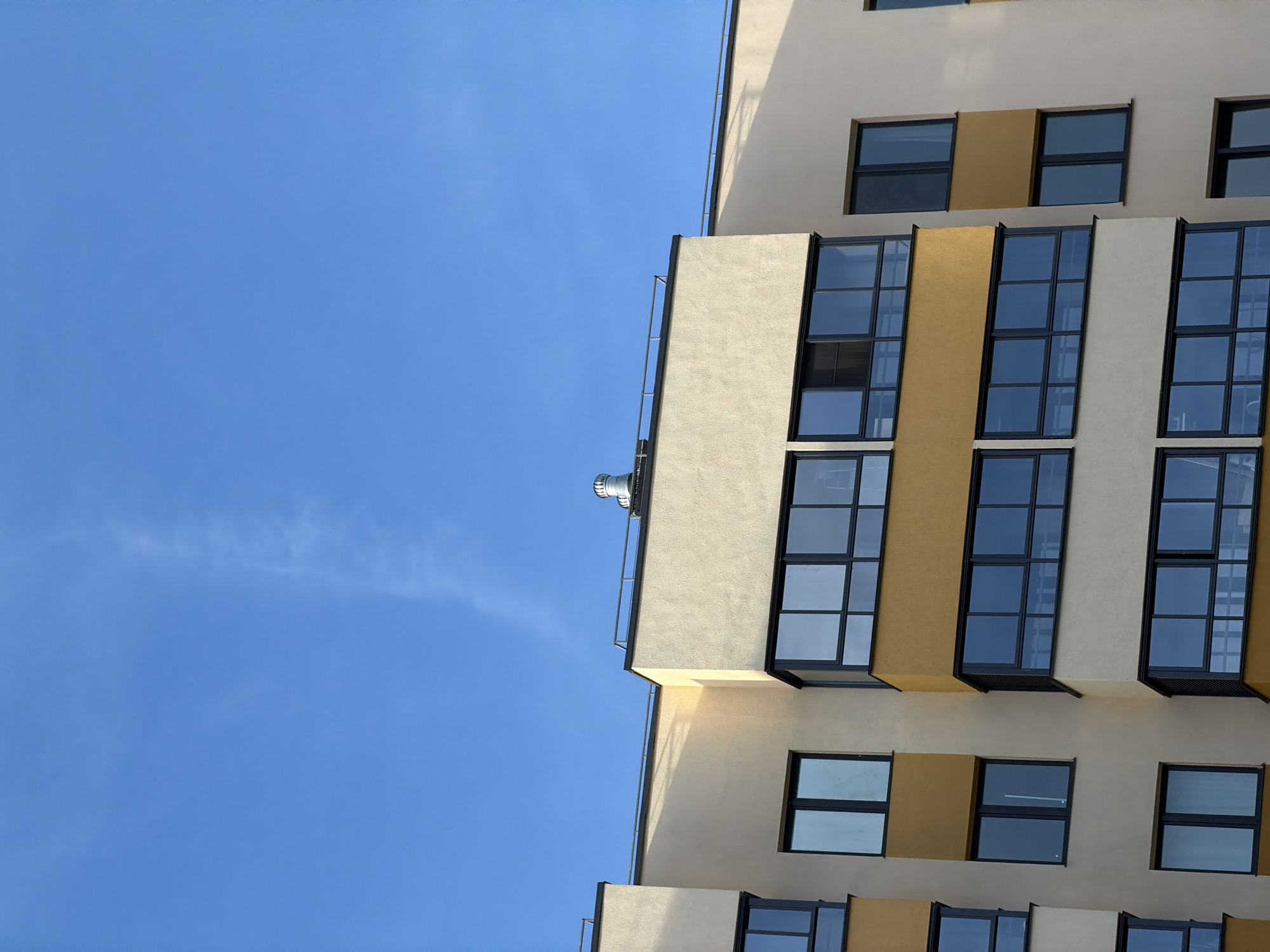 This image depicts the upper portion of a modern residential building facade under a clear blue sky in Izhevsk, Russia. The building features light cream-colored stucco sections interspersed with horizontal bands of mustard yellow. Dark-framed windows and enclosed balconies with reflective glass appear systematically across the visible floors. On the flat roofline, dark metal railings are visible, along with a single, cylindrical, metallic ventilation or exhaust fixture. Shadows on the facade suggest direct sunlight. A faint contrail is present in the upper right quadrant of the otherwise unclouded blue sky. No individuals, explicit actions, or interactions are visible in the scene.