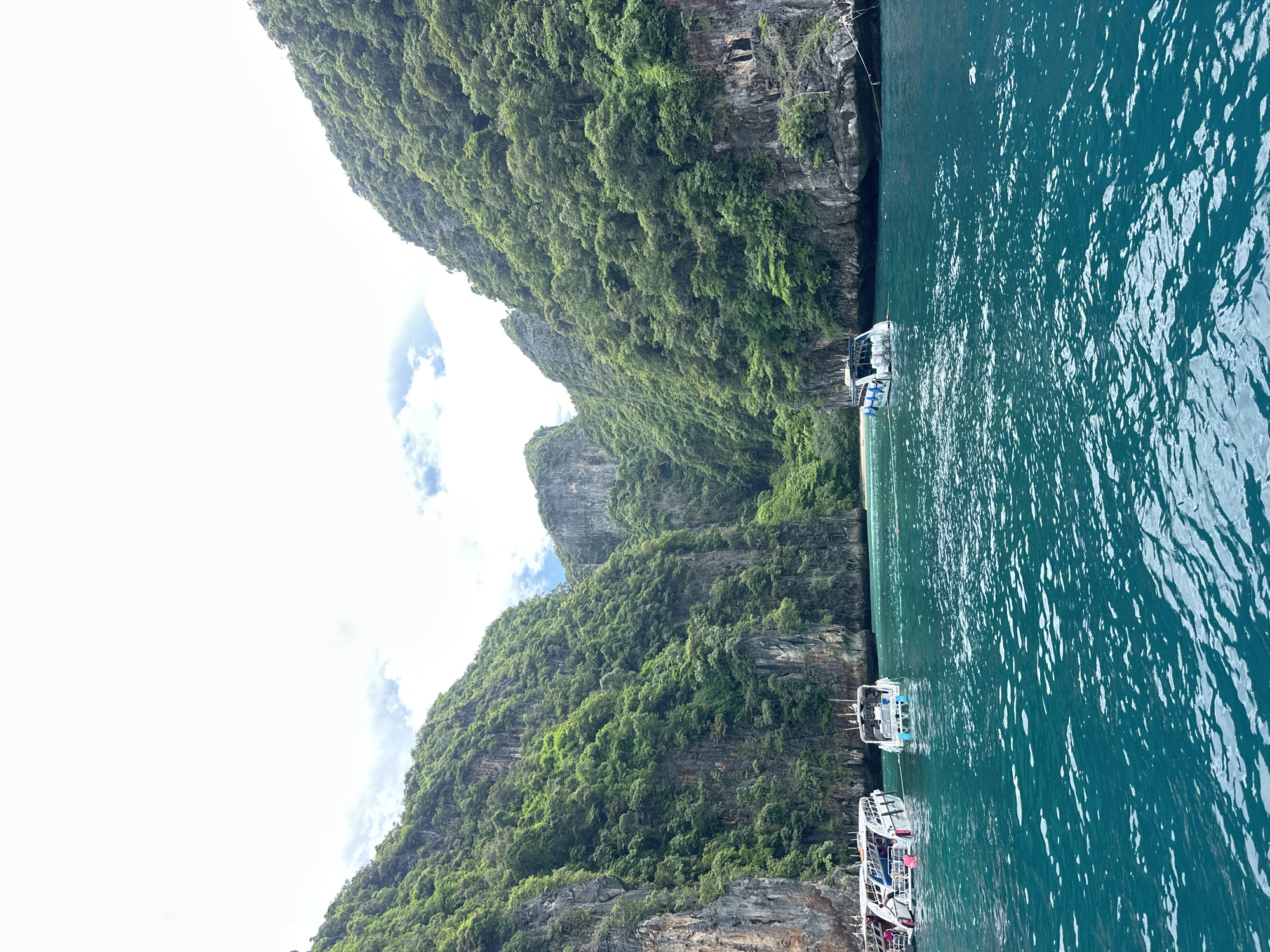A vertical, medium shot captures a view from a boat on turquoise water, looking towards a narrow bay framed by steep, lush green cliffs. Several white speedboats are anchored in the water. The sky is overcast with bright patches of white and some scattered clouds. The water is choppy with whitecaps, and sunlight reflects off its surface. The cliffs are densely covered in vegetation, with exposed rock faces visible in places. The scene depicts a popular tourist destination in the Andaman Sea, likely the Maya Bay or a similar bay near Ao Nang, Thailand, known for its dramatic limestone formations and clear waters.