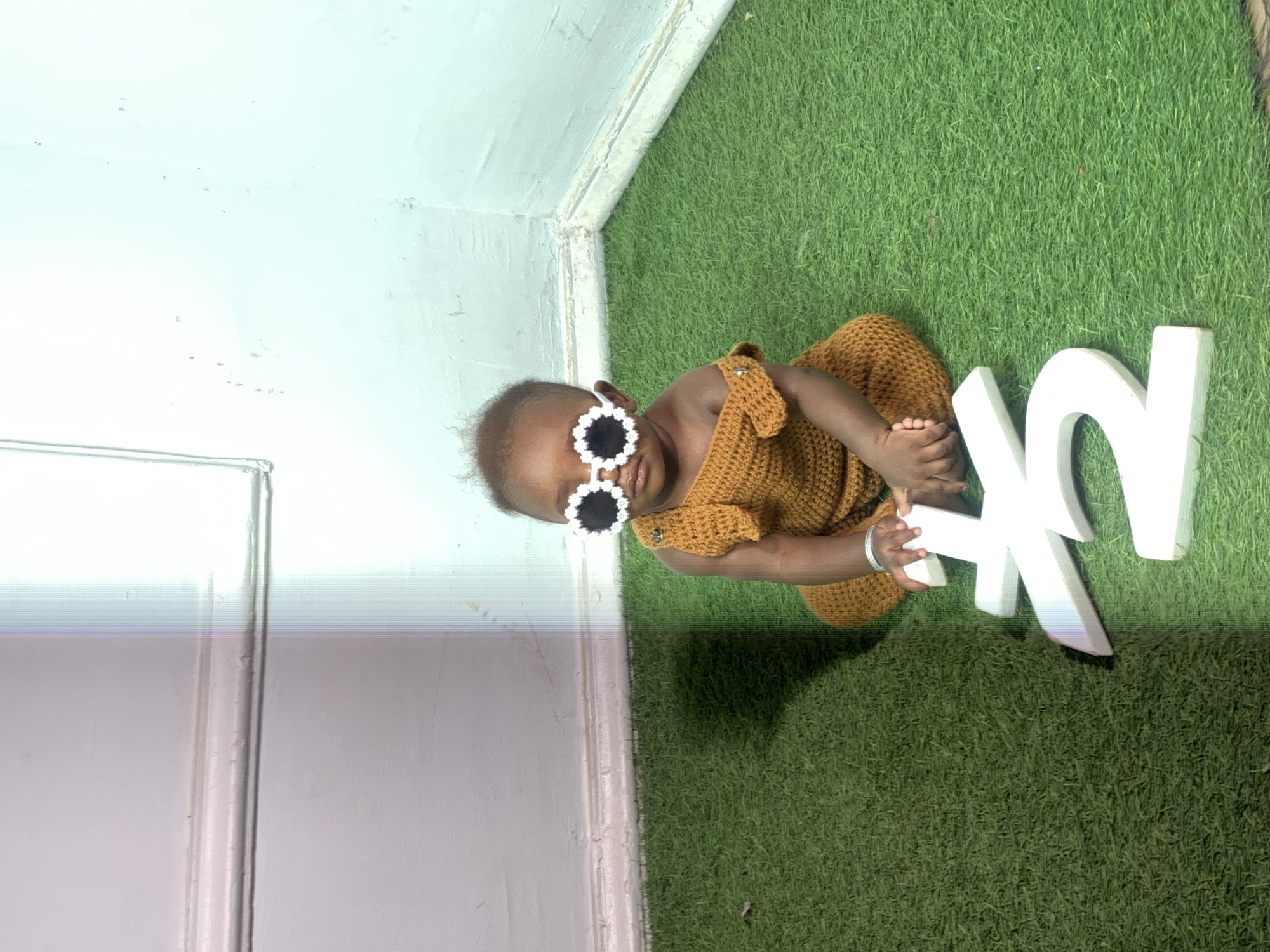 A baby with dark skin wears sunglasses and a crocheted brown outfit, sitting on artificial grass next to white block letters. The scene is indoors, near a white wall. The location is Barakin Akawo, Nigeria.