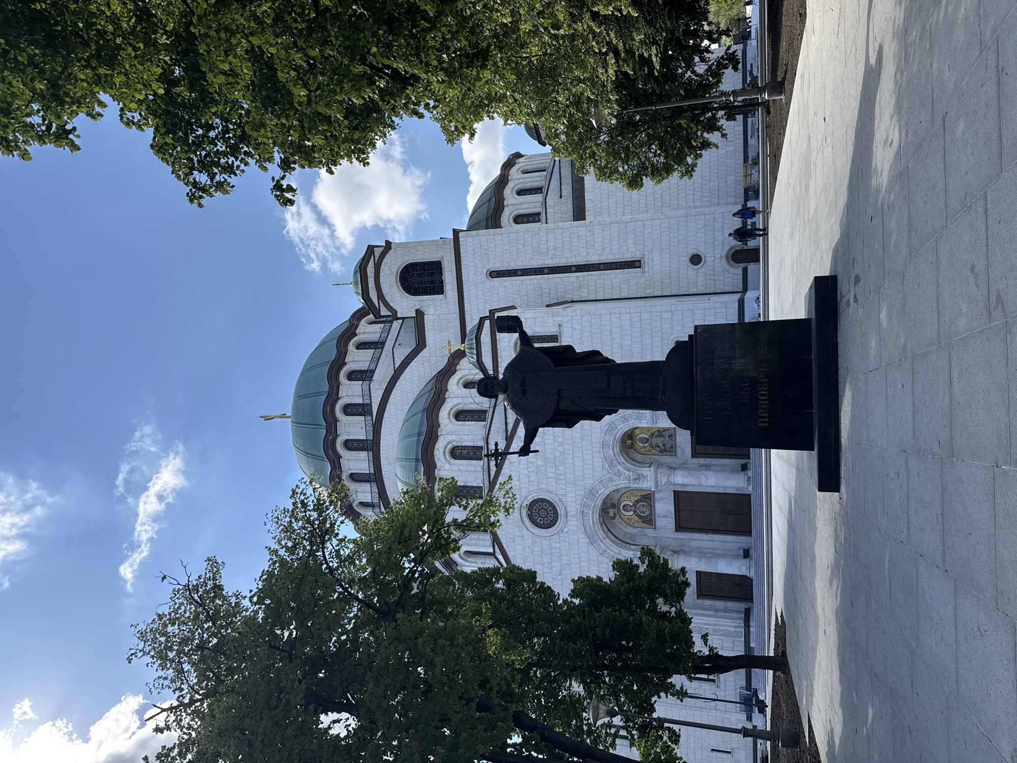 The image captures the Saint Sava Temple in Belgrade, Serbia. A bronze statue stands in the foreground on a paved plaza. The statue depicts a figure holding a cross. The temple, constructed of white stone, features a large green dome. The sky is blue with scattered clouds. Tree branches frame the scene, partially obscuring the sky and upper portion of the temple. Several people are standing in front of the temple. A lamppost is visible near the bottom right of the image. The image has been rotated 90 degrees clockwise.