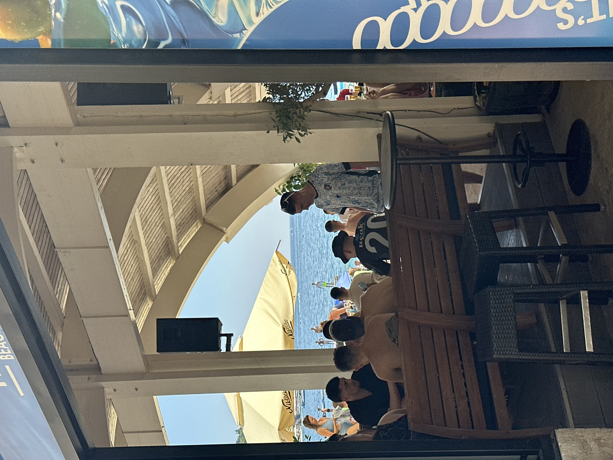 The image depicts a scene at a beach bar in Budva, Montenegro. In the foreground, several people are seated at wooden benches, facing away from the camera. One person, a young male wearing a black t-shirt and a hat with the number 20 on it, is visible on the right. To his left, two men are engaged in conversation. Further back, a young man in a patterned t-shirt and sunglasses stands near a high-top table. Beyond the seating area, beach umbrellas are set up, and people are visible on the beach and in the water. The backdrop is the Adriatic Sea under a clear blue sky. The architecture suggests an open-air establishment with supporting pillars and a slatted roof. A large blue advertisement or sign is partially visible on the far right of the image.