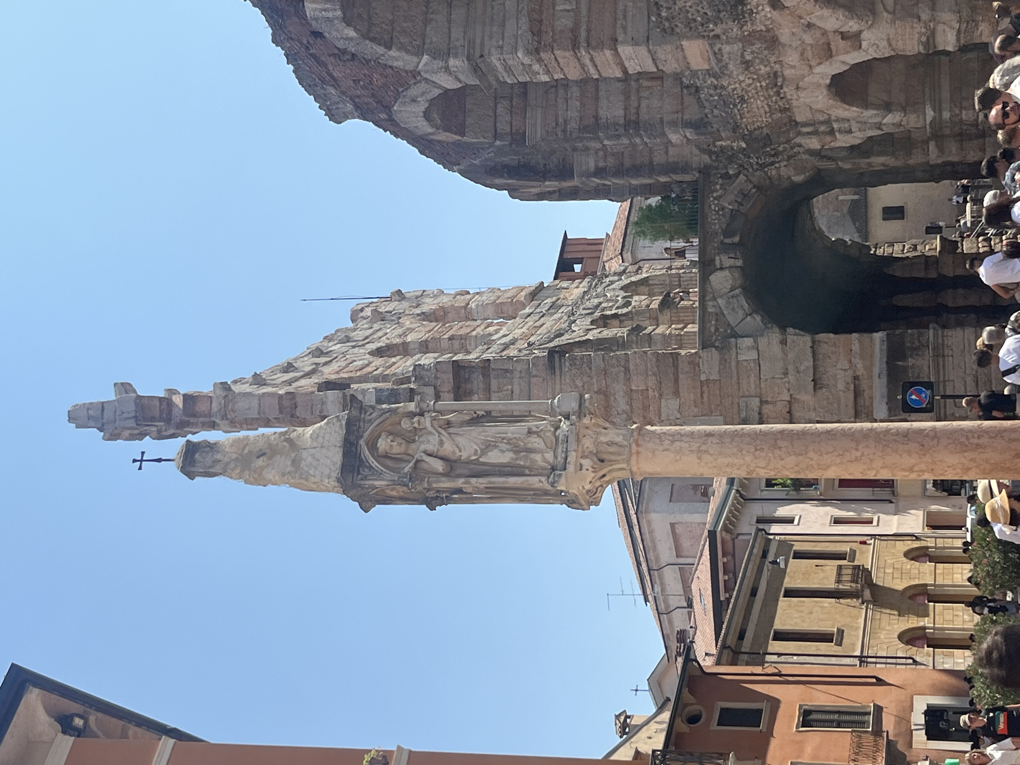The image depicts a sunny day in Piazza Bra in Verona, Italy, with a view of the Verona Arena. In the foreground, a tall, slender column with a carved sculpture of a figure holding a child is visible. Behind this, the monumental stone structure of the Arena dominates the scene, with its iconic arches and weathered facade. A cross atop a spire juts out from a section of the Arena, reaching towards the clear blue sky. Several people are scattered around the piazza, some walking, others appearing to be tourists. The architecture suggests a historical urban setting, with buildings featuring terracotta roofs and arched windows lining the periphery. The overall impression is one of a vibrant, sun-drenched public space within a historically significant Italian city.