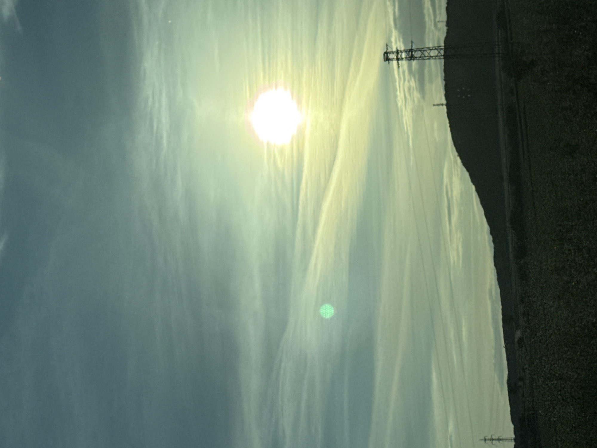 The image shows a sky with thin clouds and the sun, taken outdoors. Power lines and transmission towers are visible to the right of the frame. Adjacent to the power lines, there appears to be a hillside or elevated terrain with dense vegetation. The location cannot be confirmed as Slavyani, Bulgaria based on this image alone.
