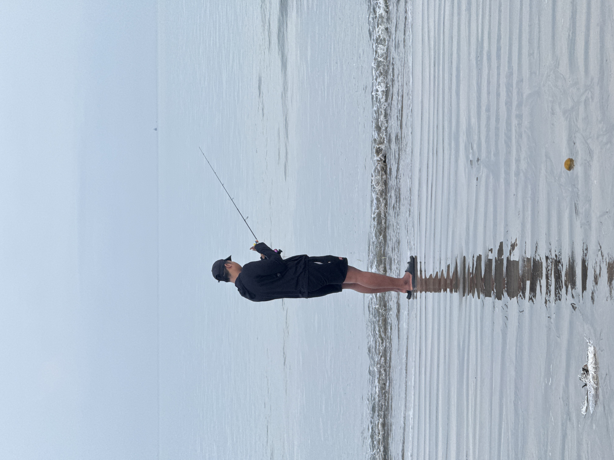 A medium shot captures a man fishing at the beach in Jimbaran, Indonesia. The sky is overcast and the air is misty. The man is positioned horizontally, facing left. He wears a dark-colored long-sleeved shirt, shorts, and a baseball cap. He also wears sandals and holds a fishing rod, its line extending into the sea. The beach has visible water ripples, and there is a small, round, yellowish object on the sand to the right. The man's reflection is mirrored in the wet sand.