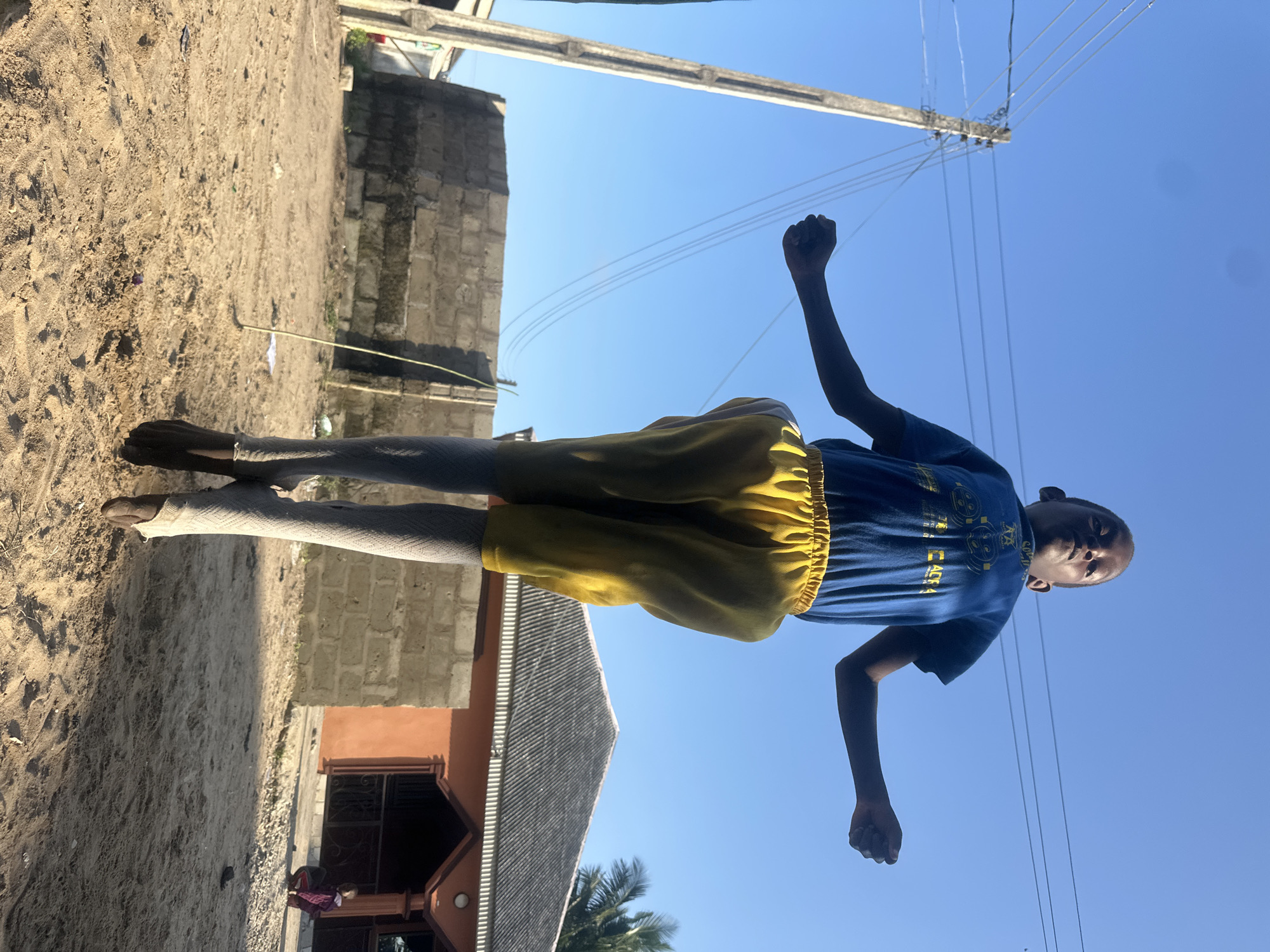The media file shows a young person in Badagry, Nigeria, standing outside under a clear blue sky. The person is wearing a blue t-shirt with yellow text and graphics, and yellow shorts over grey leggings. Their right arm is raised in a fist, suggesting a gesture of strength or defiance.
The setting appears to be a sandy area with a partially constructed building made of concrete blocks in the background. To the right, electrical wires are visible against the sky, strung from a utility pole. In the lower part of the image, a building with an orange facade and a dark-tiled roof is partially visible, with a glimpse of another person in a purple garment standing near the entrance.
The lighting suggests it is daytime, likely morning or afternoon, given the bright sunlight. The overall mood is somewhat energetic, perhaps indicating a moment of play or a posed photograph. The text on the t-shirt is partially obscured but appears to have some Japanese characters and other symbols.