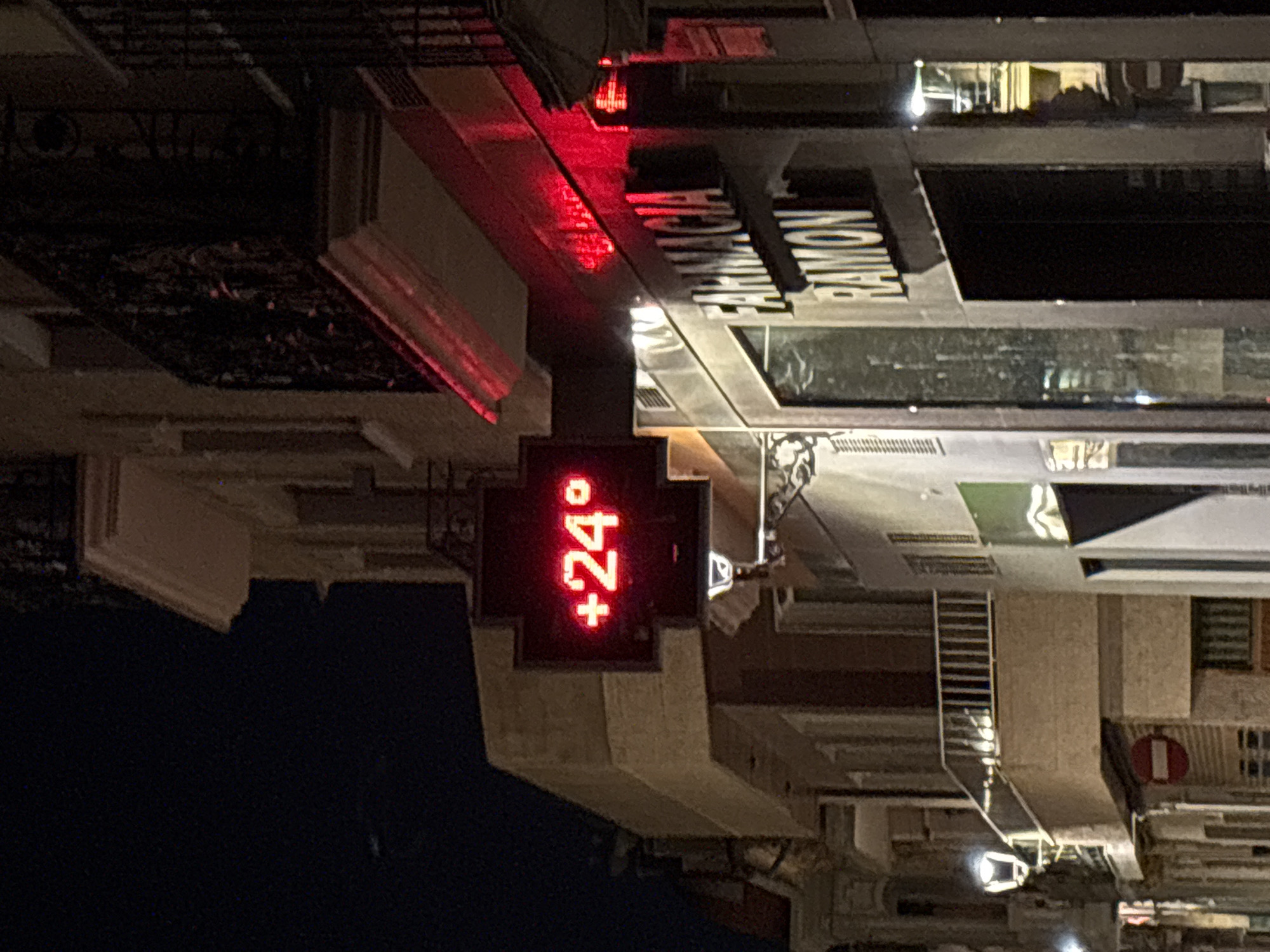 The image shows a street scene at night in Picassent, Spain. A red cross-shaped thermometer displays a temperature of "+24°". Above it, a building facade is visible with balconies and ornate railings. To the right, a pharmacy sign reads "FARMACIA RAMON" in white capital letters on a dark background. The street is illuminated by streetlights and the red glow of the thermometer, casting shadows on the buildings and pavement. No people are visible in the frame.