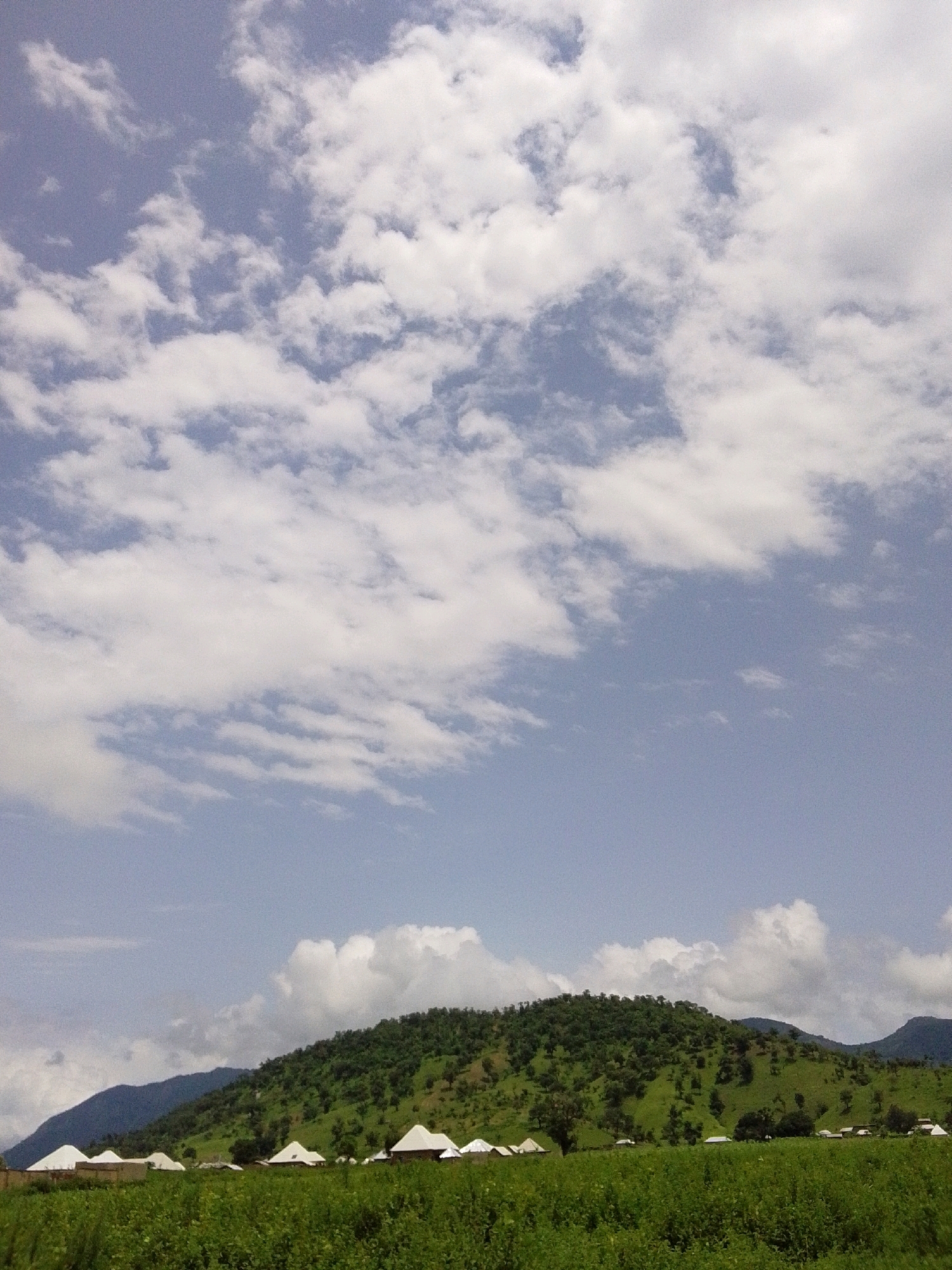 The image depicts a rural landscape in Serti, Nigeria, under a sky filled with scattered white clouds against a pale blue backdrop. In the foreground, a field of lush green vegetation stretches across the bottom of the frame. Beyond the field, a cluster of buildings with conical, white roofs are visible. These structures are set against the backdrop of a verdant, rolling hill that rises behind them. The hill is densely covered with trees and scrub, with darker, more distant mountains forming the horizon. The overall impression is of a peaceful, natural setting, typical of rural Nigerian countryside.