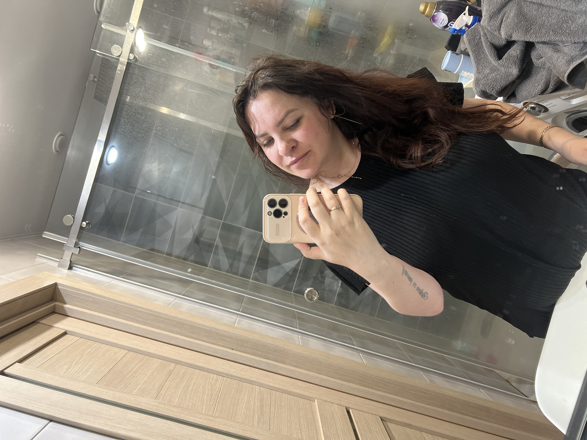 A woman takes a selfie in a bathroom with a glass shower door and light wood paneled walls. A washing machine is visible in the background. No information is available to determine if the bathroom is in Noviy, Russia.