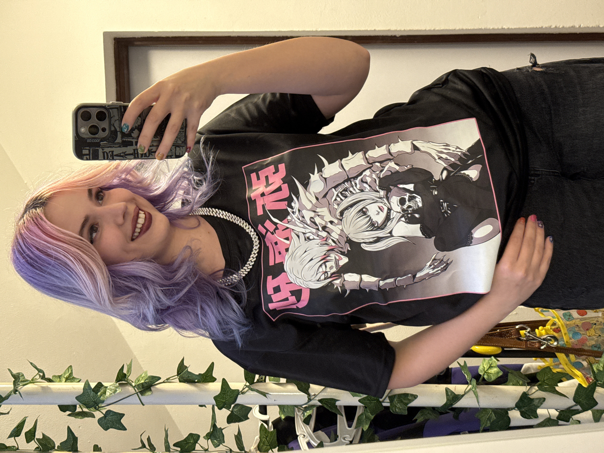 A fair-skinned female with pink and purple hair is holding a smartphone up to take a selfie. She is smiling, wearing a silver chain necklace, and a black T-shirt with a manga design in a white and pink bordered rectangle. She also wears black jeans with a hole in the thigh. In the background is a mirror and a light wall. Faux ivy is wrapped around a white object. The location is not determinable from the image.