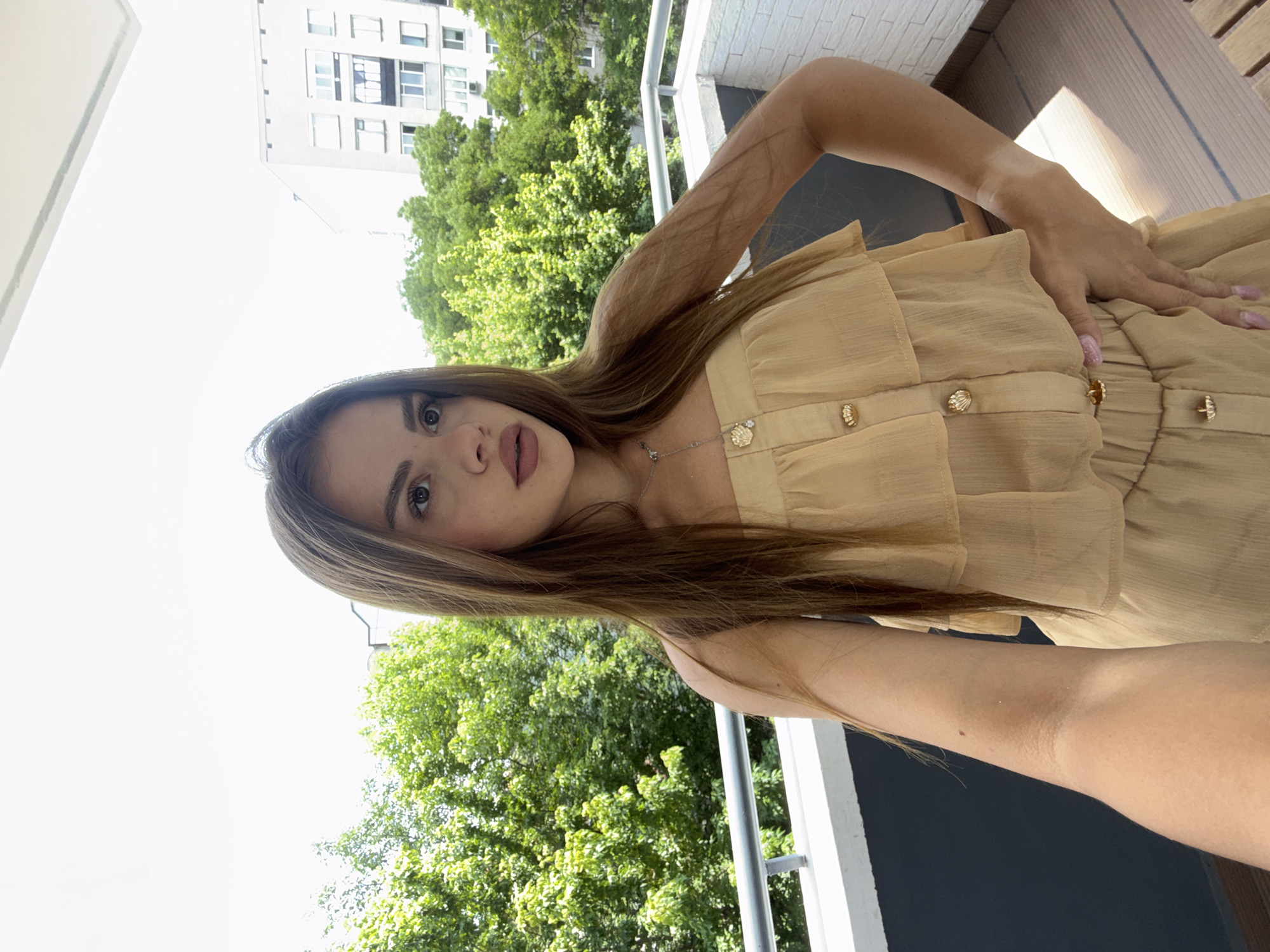 This vertical photograph, taken in Plovdiv, Bulgaria, captures a young woman posing outdoors on a balcony or terrace during daylight hours. The woman, with long, straight brown hair, looks directly at the camera with a neutral expression. She has light-colored eyes and wears light makeup, including a light-toned lipstick on her full lips.
She is dressed in a light beige or tan strapless top or dress featuring ruffled details on the bodice and several ornate gold-colored buttons, resembling seashells, running down the front. A delicate silver necklace with a small pendant is visible around her neck. Her nails are painted a light lavender or pink. Her right arm, visible in the foreground, shows a distinct tan line.
The background features vibrant green trees, suggesting a verdant environment. Above the trees, a large, light-colored, multi-story building, likely an apartment complex, is visible against a bright, partly overcast or overexposed sky. A white metal railing, consistent with a balcony setting, runs along the right side of the frame. The bright illumination indicates a sunny day. There is no visible text in the image.