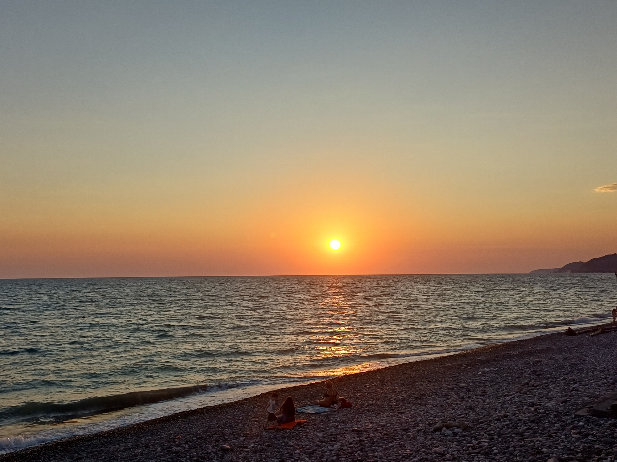 A tranquil sunset unfolds over a pebble beach in Sochi, Russia. The sun, a vibrant orb, casts a golden glow across the rippling sea, its reflection creating a shimmering path on the water's surface. The sky transitions from a soft blue at the top to a warm orange and yellow near the horizon, indicating the end of the day.
On the beach, a few individuals are enjoying the serene atmosphere. In the foreground, three people are gathered, one sitting on a towel and two others nearby. Further down the beach, another person is visible near the water's edge. The beach itself is covered in smooth, grey pebbles. The gentle waves lapping at the shore add to the peaceful ambiance. In the distance, a hilly coastline is faintly visible against the horizon. The overall mood is one of relaxation and appreciation for the natural beauty of the sunset. There is no visible text in the image.