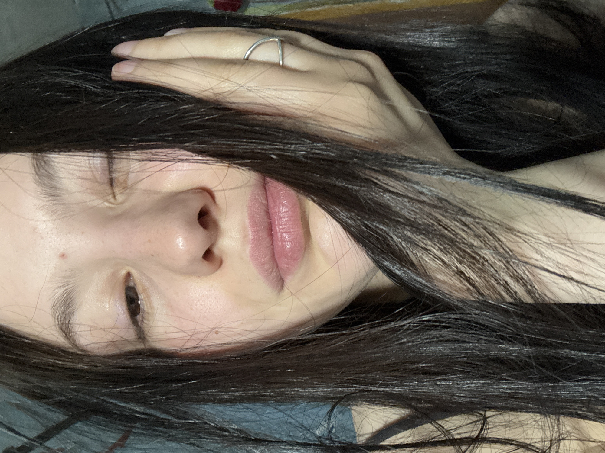 A close-up, slightly angled shot depicts a person with long, dark hair partially obscuring their face. The person is lying down, with their hand resting on their head. A silver ring is visible on one of the fingers of the hand on their head. The person's eyes are partially closed, and their lips are a light pink color. The background is blurry and indistinct.
There is no information or indication that the photo was taken in the city of Фоки, Russia.
