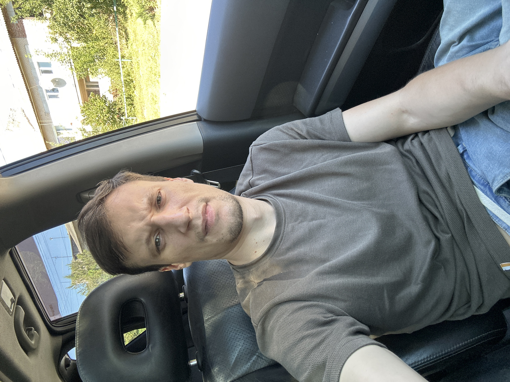 A medium shot shows a light-skinned man with short brown hair and stubble sitting in the driver's seat of a car. He is wearing a brown t-shirt and blue jeans. The man is looking directly at the camera with a neutral expression. The interior of the car is visible, including the black leather seat and the gray dashboard. The windows are open, and an outdoor scene is visible in the background. The scene appears to be in a residential area with houses and trees. The image is taken during the daytime, with bright sunlight casting shadows. The car is parked on a street or driveway. The specific location is Makiv, Ukraine, as indicated by the surrounding architecture.
