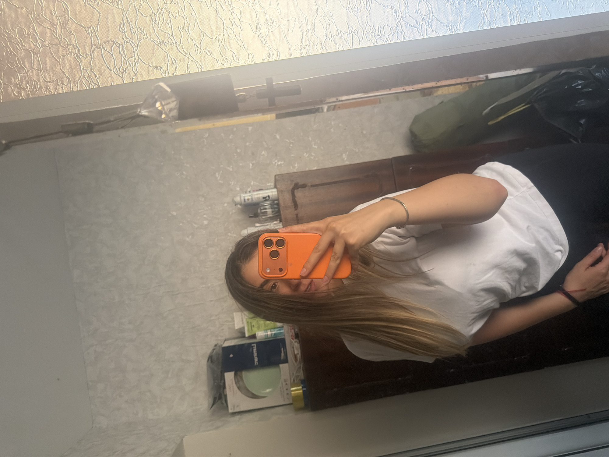 A young woman is depicted taking a mirror selfie, holding an orange smartphone in her right hand, which partially covers her face. She has long, light brown hair flowing over her shoulders and is wearing a white t-shirt and dark pants. A thin silver bracelet adorns her right wrist, while her left wrist bears a dark band with a red accent.
The selfie captures an indoor scene reflected in a large mirror. The background wall in the reflection features a light-colored, textured wallpaper with a subtle pattern. Below the reflection of the woman, a wooden surface, possibly a dresser or shelf, holds various items. These include a white spray can with visible text "R." and a dark blue box displaying "TUMAC" and "PROFESS" in white lettering, alongside other small containers. To the right, a dark green object, resembling a rolled-up mat or bag, and a black plastic bag are visible.
Above the reflected room, a window or translucent panel with a patterned glass texture suggests the presence of natural light. A small, reflective metallic object is visible in the upper left of the reflected image, and a dark, cross-shaped fixture appears in the upper right. The image is oriented sideways, with the subject appearing horizontally.