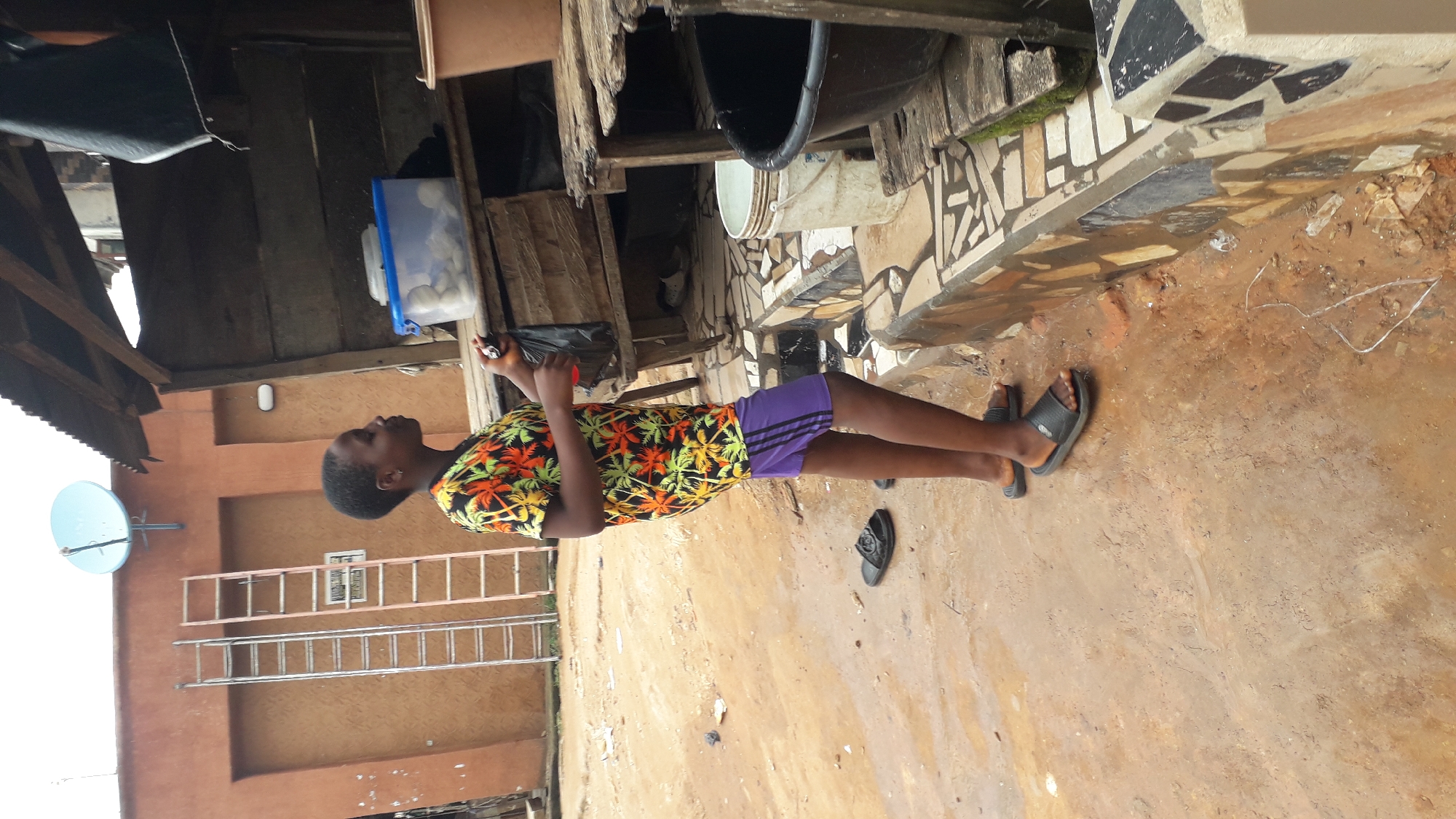A young person stands on a dirt ground in Nkpor, Nigeria, wearing a colorful, patterned short-sleeved shirt and purple shorts with black stripes. They are facing right, holding a black plastic bag and a red bottle in their hands. The person is wearing black flip-flops. To the left of the person, two ladders lean against an orange-brown wall. A satellite dish is mounted above the ladders on the wall. Behind the person, a rustic wooden structure with shelves is visible, containing a clear plastic container filled with round white objects. Various containers and buckets are also present around the structure. The ground is wet and appears to be composed of mud and dirt, with some scattered debris. The environment suggests an outdoor, possibly commercial or residential, setting in an urban area.