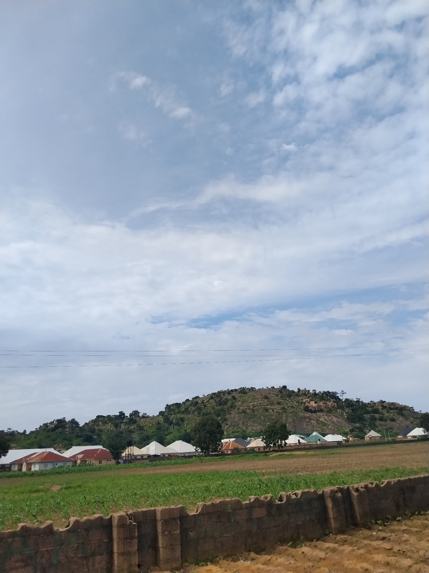 The image captures a view of Nabatum, Nigeria, under a cloudy sky. In the foreground, a rough, textured brown brick wall runs horizontally across the frame. Beyond the wall lies a cultivated field of green crops, possibly maize, which appears to be growing vigorously. In the middle ground, a cluster of houses with various roof colors, including red, white, and green, are situated. These homes are nestled against a backdrop of a rocky, tree-covered hill that dominates the upper portion of the image. The overall scene suggests a rural or semi-rural setting within the city.