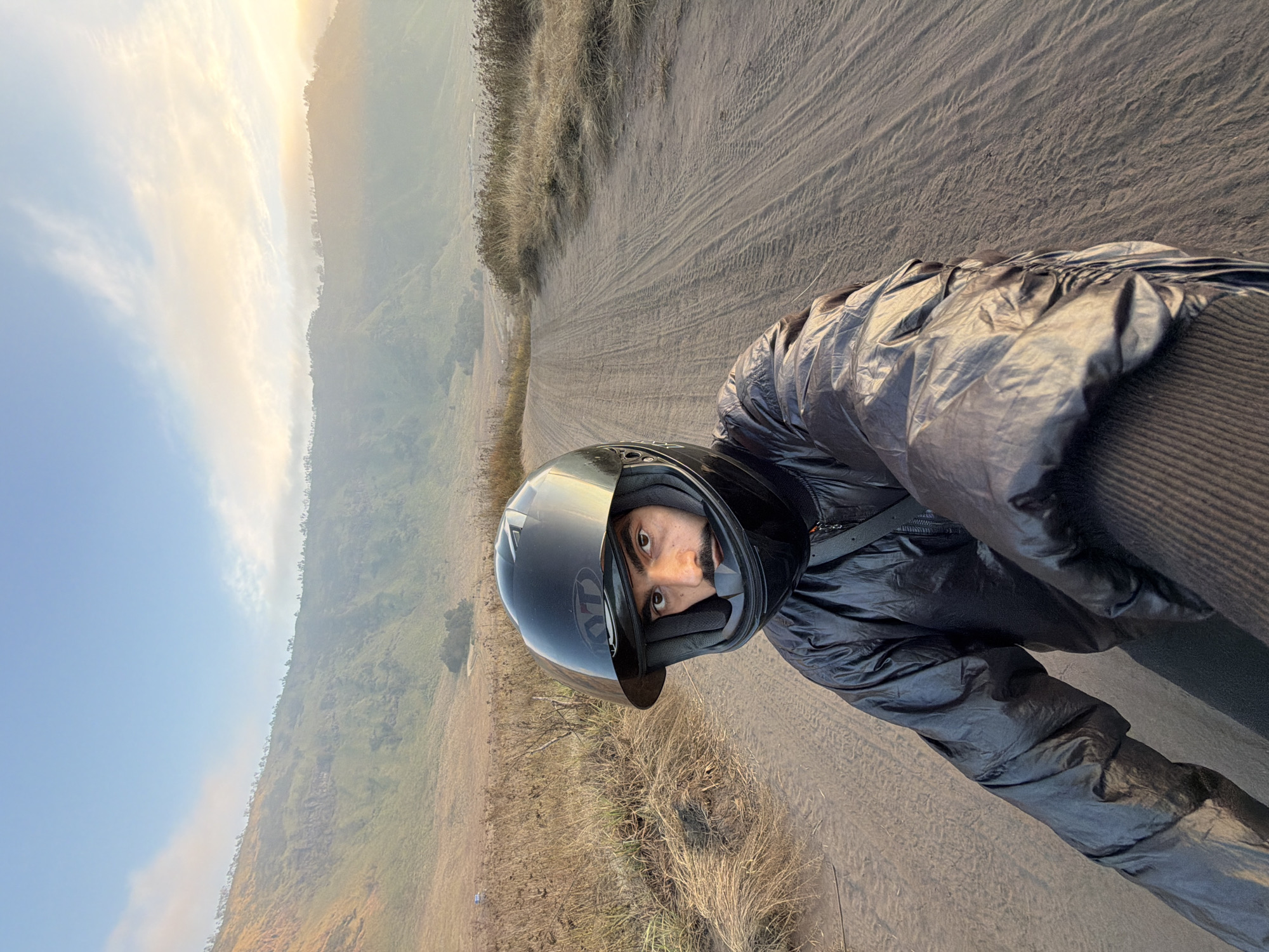 An overhead, first-person perspective selfie shows a man wearing a black motorcycle helmet and a black puffer jacket. He is looking directly at the camera with a neutral expression. The man is positioned on a dirt road that stretches into the distance. To the left and right of the road are dry, brown grasses and the lower slopes of a green, mountainous landscape. The sky above is a clear blue with some wispy clouds, tinged with the orange and yellow hues of a sunrise or sunset. The overall scene suggests an outdoor adventure in a natural, possibly volcanic, environment. The location is likely Bromo Tengger Semeru National Park, East Java, Indonesia, given the distinctive landscape.