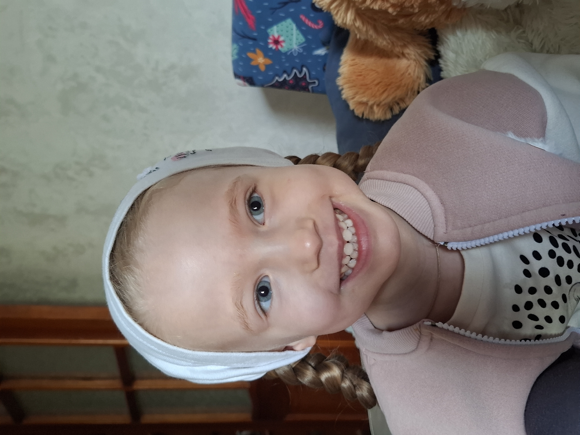 A portrait of a young fair-skinned girl with blue eyes, light blonde hair braided into two pigtails, wearing a white headband, a pink sweater over a polka-dot shirt, smiling widely, facing the camera. The background includes a blue patterned object and a brown plush toy. The image was likely taken indoors. The exact location in Chaikovsky, Russia cannot be determined from the image alone.