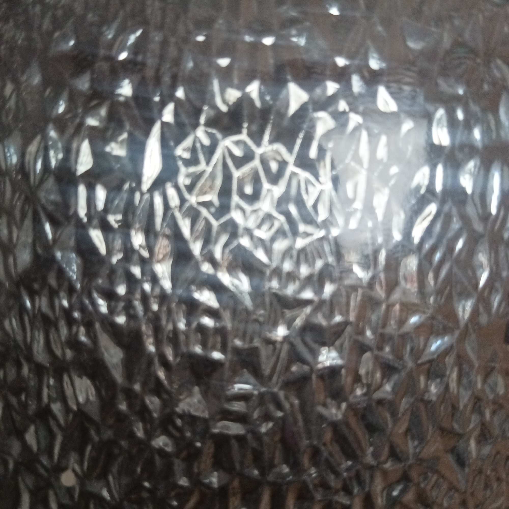 The image depicts a textured glass surface. The glass features a repeating pattern of geometric shapes, including triangular and hexagonal forms, which create a distorted and reflective effect. The color palette is monochromatic, ranging from dark gray to bright white due to the light reflecting off the glass facets. No people, subjects, objects, actions, or interactions are visible. The environment is limited to the immediate surface of the glass. There is no information in the image to support the assertion that this image was taken in Chaikovsky, Russia.