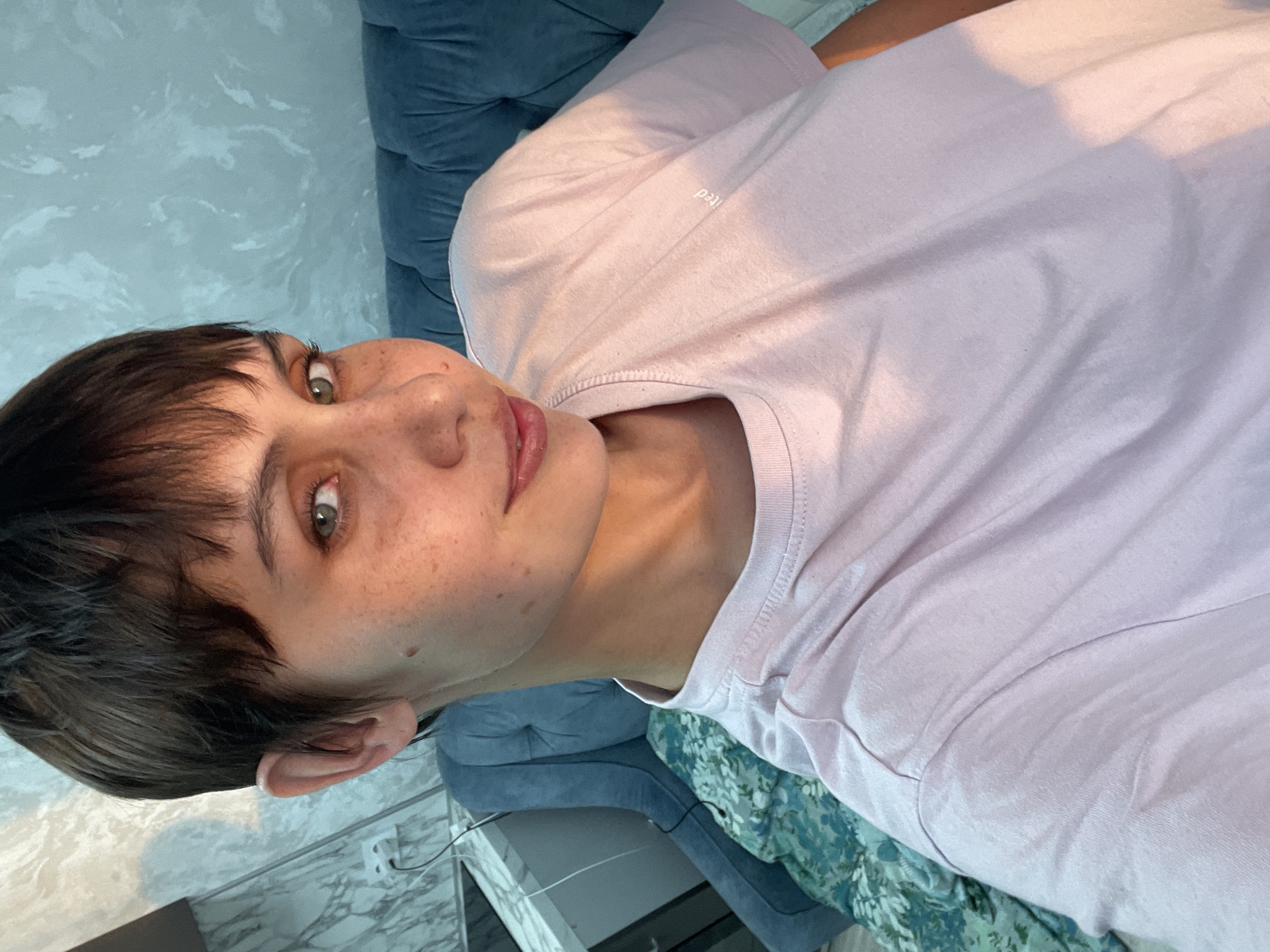 A close-up, portrait-oriented image shows a young person with short, dark brown hair and fair skin. They have freckles across their nose and cheeks and green eyes. The person is wearing a light pink t-shirt. They appear to be sitting on or leaning against a blue tufted piece of furniture, possibly a couch or headboard. Part of a wall with a textured pattern is visible behind the furniture. A floral patterned fabric can be seen below the blue tufted surface. There's a white, reflective surface and a glimpse of electrical wiring in the bottom left corner. The location, Izhevsk, Russia, is not discernible from the image alone.