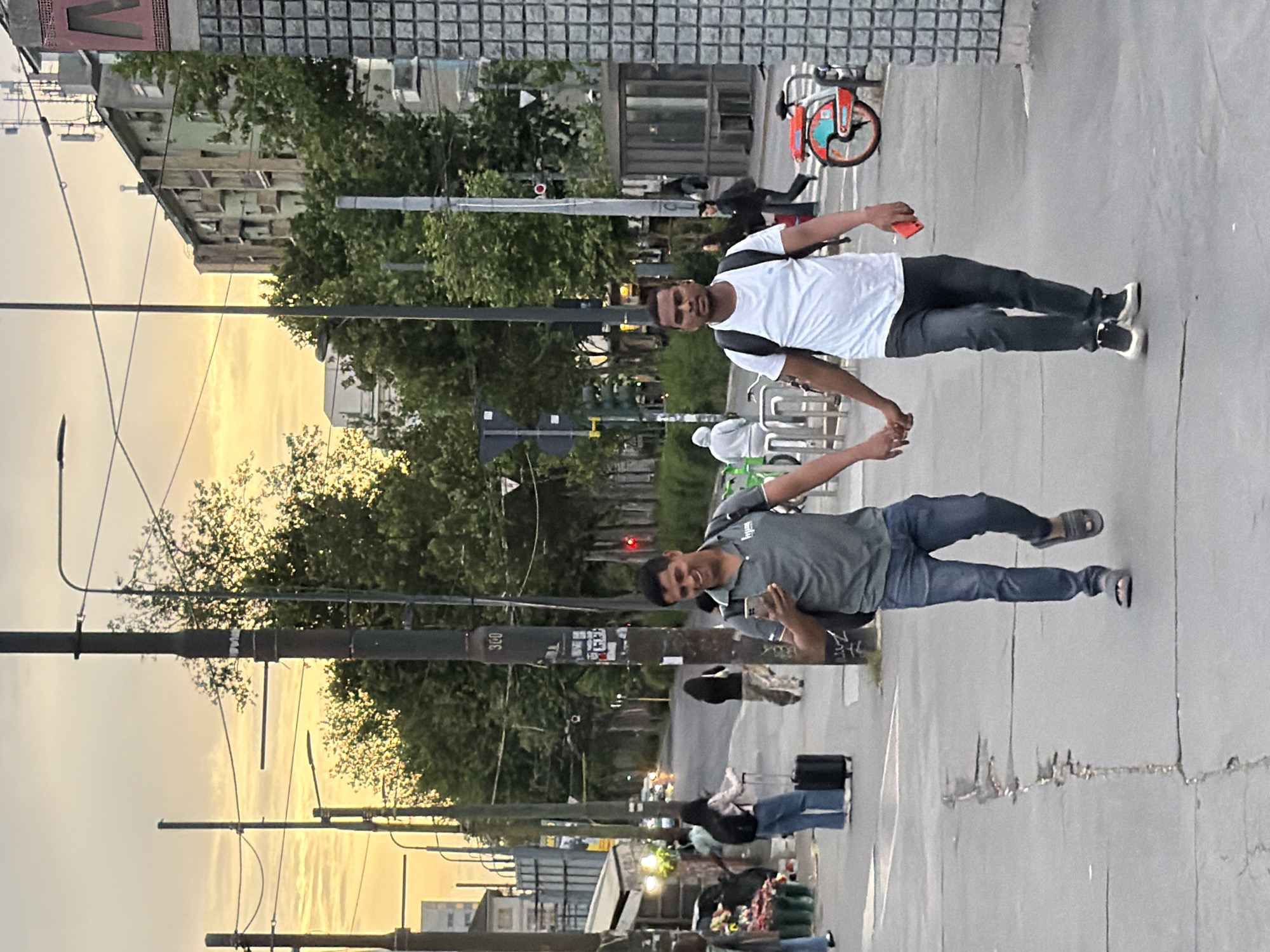 The image shows two men holding hands on a sidewalk in Milan, Italy. The man on the right wears a white t-shirt, black pants, and a backpack, while holding a red phone. The man on the left wears a gray t-shirt, jeans, and sandals, while looking at his phone. The background includes buildings, trees, streetlights, a bicycle, and other pedestrians. The time of day appears to be late afternoon or early evening, with a sunset visible in the background.