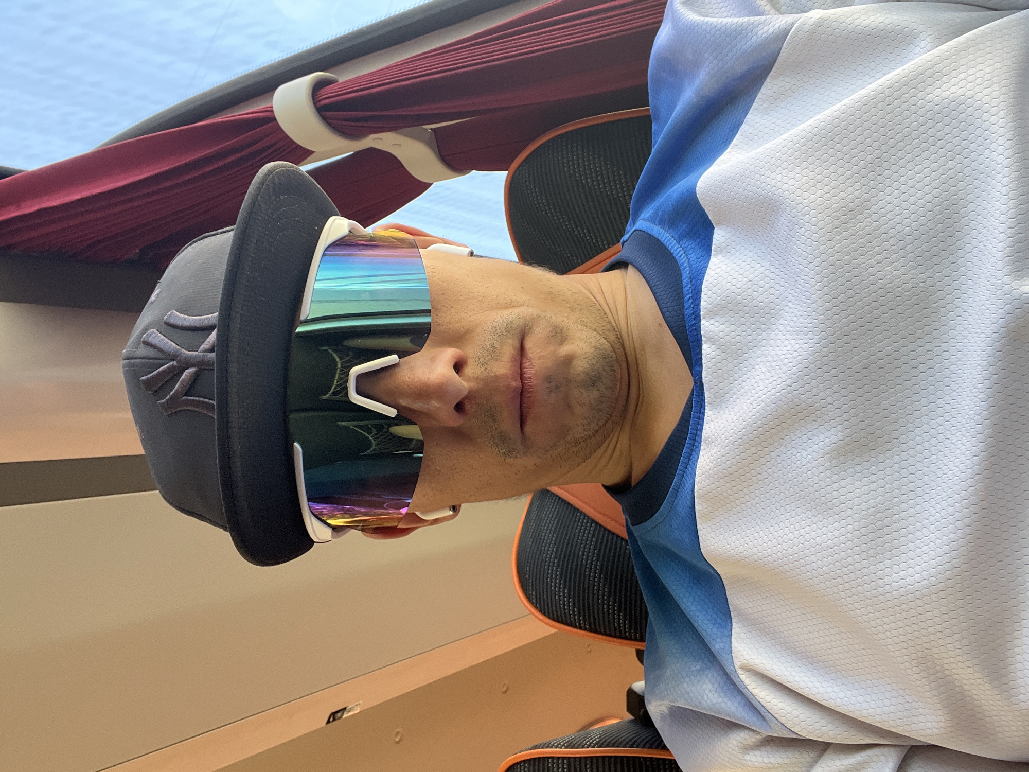 A man wearing a baseball cap, sunglasses, and a white and blue jersey is photographed from below. The cap is dark gray with a logo on the front. The sunglasses have iridescent lenses and white frames. The man's face is partially obscured by the sunglasses. Above him, there is a red curtain and a window, suggesting an indoor setting, possibly in Chaikovsky, Russia. There is also a chair behind him. The photograph appears to have been taken inside a vehicle.