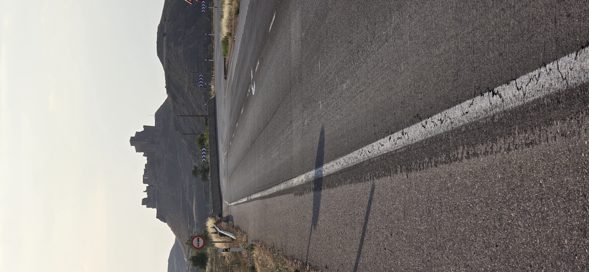 This media file captures a view of a road with a mountain range in the background. The road is paved with asphalt and has a white line marking its edge. There are several traffic signs visible along the roadside, including a red circular sign with a horizontal bar, which typically indicates a prohibition. The mountain on the left side of the image appears to have some structures or ruins built on it, giving it a historical or ancient appearance. The sky is bright and appears to be clear, suggesting it is daytime, possibly late afternoon or early morning due to the shadows cast on the road. The overall scene depicts a somewhat desolate or rural landscape, common in parts of Spain. No people are visible in the image.
