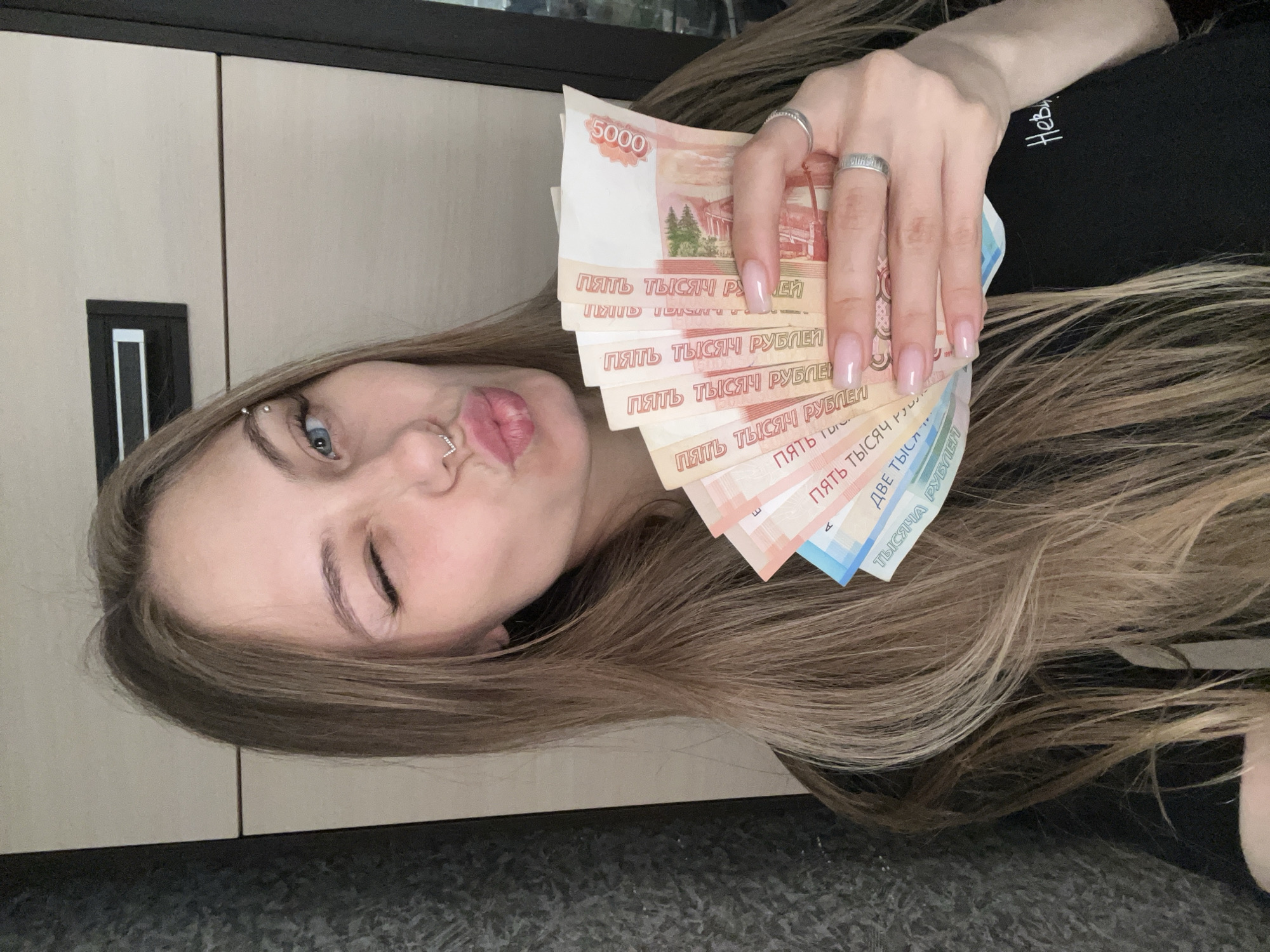 A woman is pictured holding a fan of Russian banknotes, primarily 5000-ruble notes. She is winking, has a nose ring, and wears rings on her fingers. Her hair is light brown. The background includes a light-colored cabinet and a black shirt or garment. The location is potentially Фоки, Russia, inferred from the banknotes' currency.