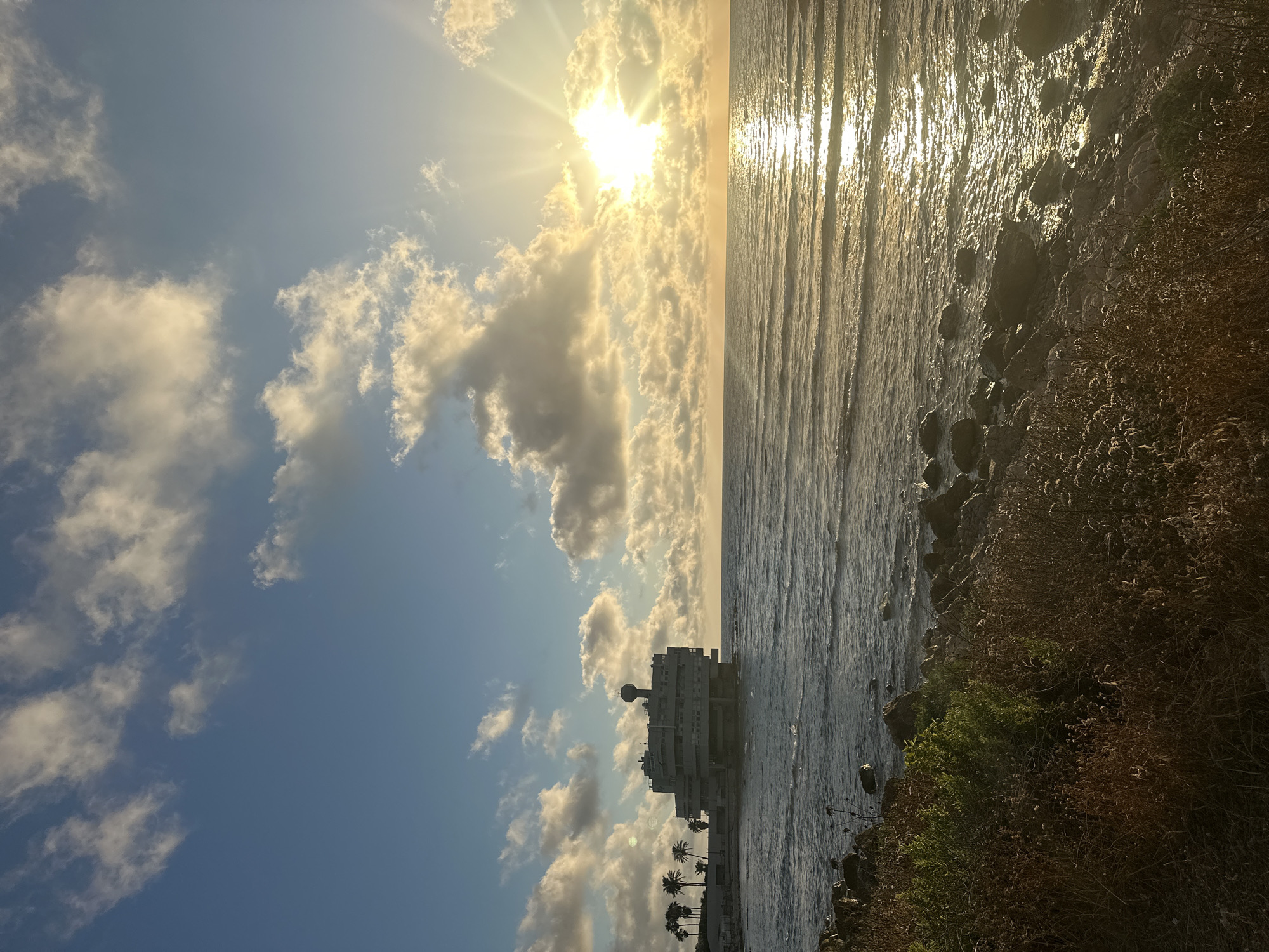 The image depicts a coastal scene in Haifa, Israel, at sunset. The sun is low on the horizon, casting a warm glow and creating shimmering reflections on the water's surface. Wispy clouds are scattered across a clear blue sky, illuminated by the setting sun.
The foreground features a rocky shoreline with dry brush and some greenery. To the left, a large, multi-story building with a distinctive dome-like structure on its roof stands near the water's edge. Several palm trees are visible in front of the building, suggesting a warm climate. The sea appears calm with gentle waves lapping at the shore.
The overall mood is serene and tranquil, capturing the beauty of a sunset by the sea. The vibrant colors of the sky and water, combined with the silhouette of the building and palm trees, create a picturesque and peaceful atmosphere. There is no visible text in the image.
