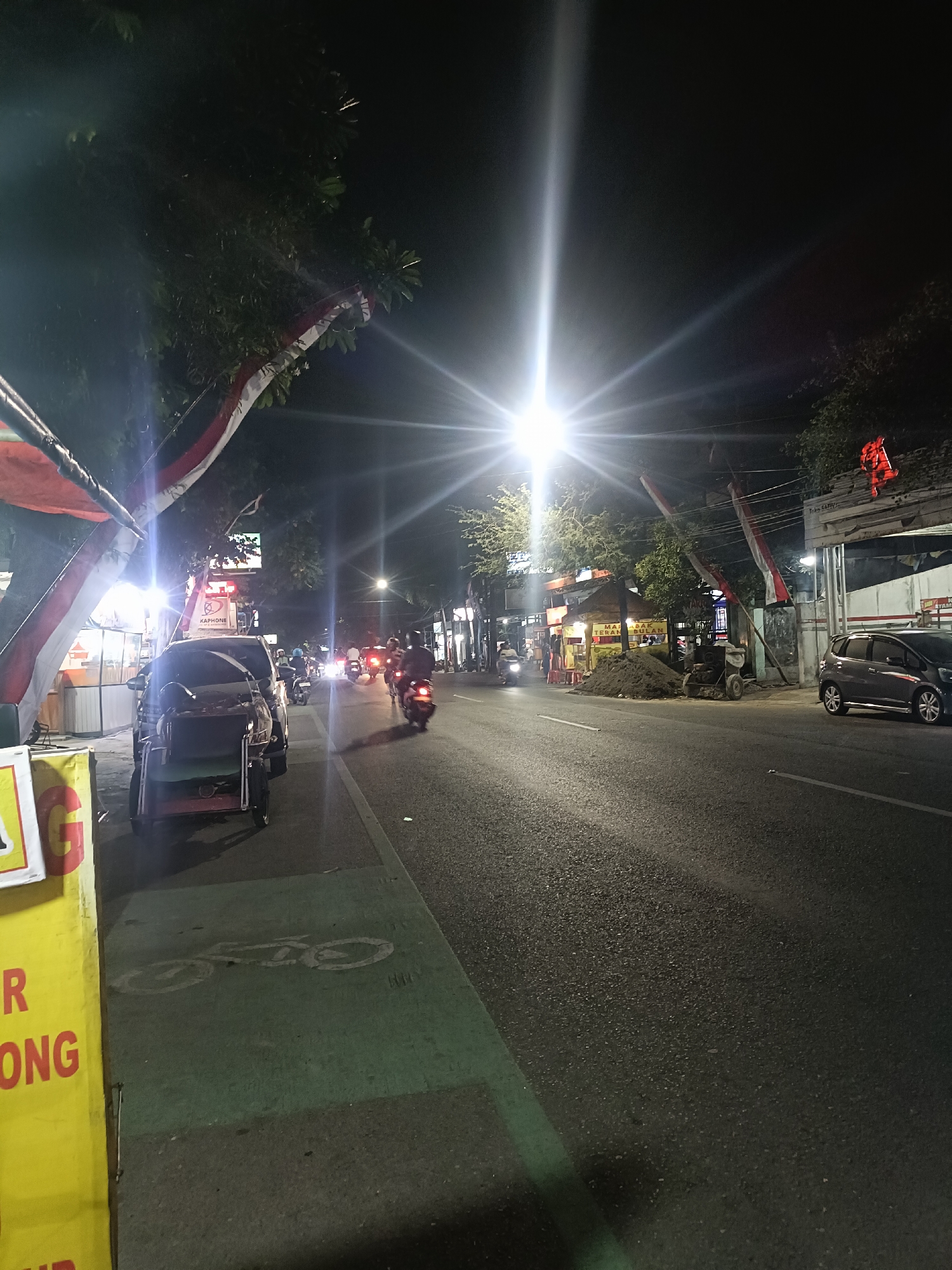 A street scene at night in Kediri City, Indonesia, is depicted. The image captures a road with several motorcycles and a car. The street is illuminated by bright lights, possibly from streetlights and businesses along the roadside. On the left side of the road, there are some food stalls or shops with illuminated signs. A yellow sign with red lettering is prominently visible. A three-wheeled vehicle, possibly a pedicab, is parked on the side of the road. The road surface is asphalt and appears to be wet in some areas, reflecting the lights. There are trees and flags, some appearing to be Indonesian flags, decorating the roadside. The overall atmosphere suggests a busy urban environment at night.