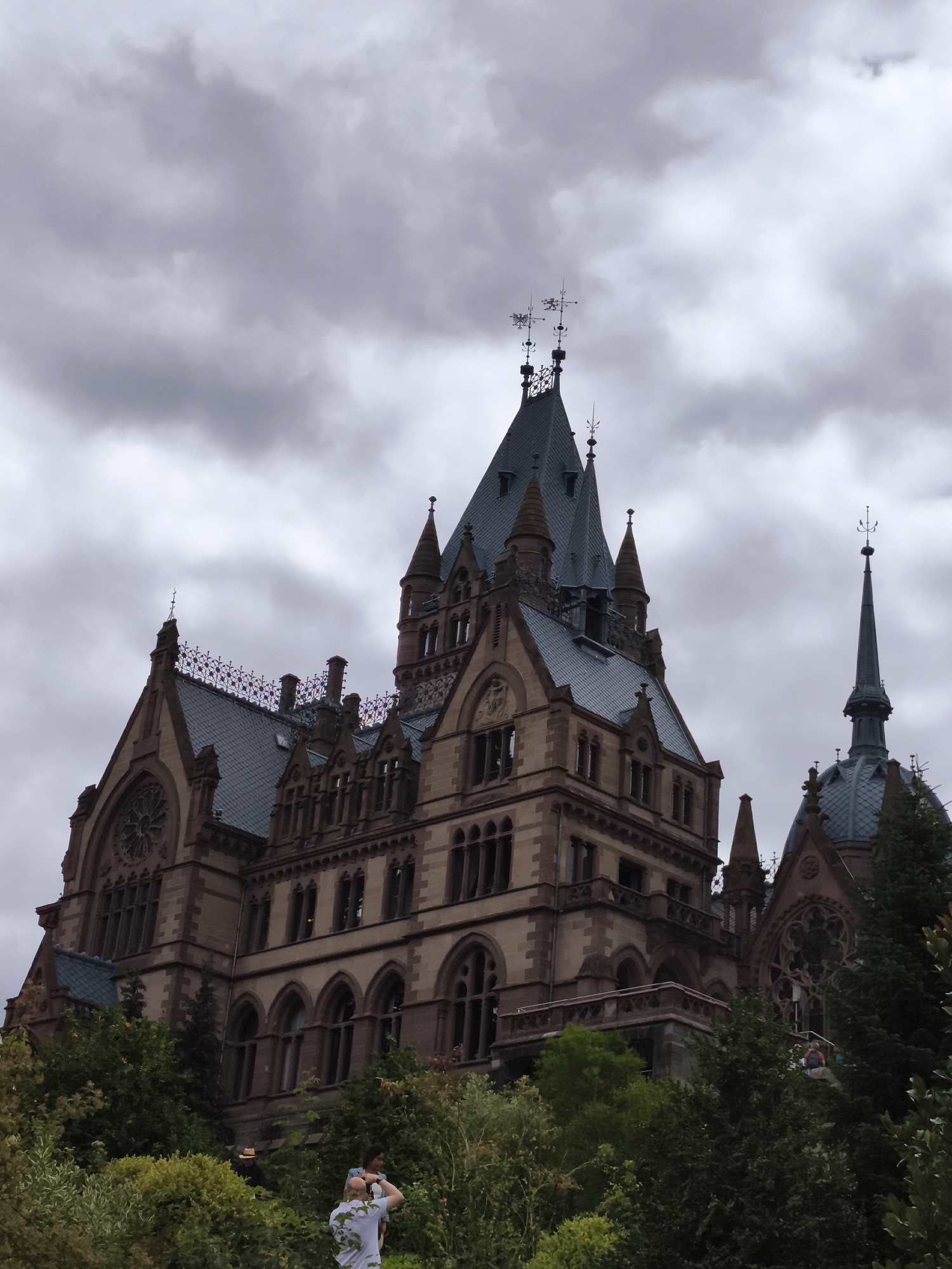 The image captures a low-angle, upward view of a grand, historic building, likely the Drachenburg Castle, situated in Königswinter, Germany. The structure is characterized by its elaborate Gothic Revival architecture, featuring multiple spires, steep roofs, arched windows, and intricate stonework. The building's facade is constructed from reddish-brown stone, contrasting with the dark grey slate roofs. Above the main structure, several ornate weather vanes adorn the spires.
The sky is overcast with heavy, grey clouds, creating a dramatic and moody atmosphere. In the foreground, lush green foliage, including trees and shrubs, partially obscures the lower sections of the building. A few individuals are visible amidst the greenery. In the lower right portion of the image, a person is partially visible, facing away from the camera and appearing to be taking a photograph with a mobile phone. In the lower center, two individuals are standing, one with their back to the camera, and the other, a woman, is wearing a backpack. Another person, wearing a straw hat, is also visible in the foliage. The overall scene depicts a picturesque view of a historic castle surrounded by nature under a cloudy sky.