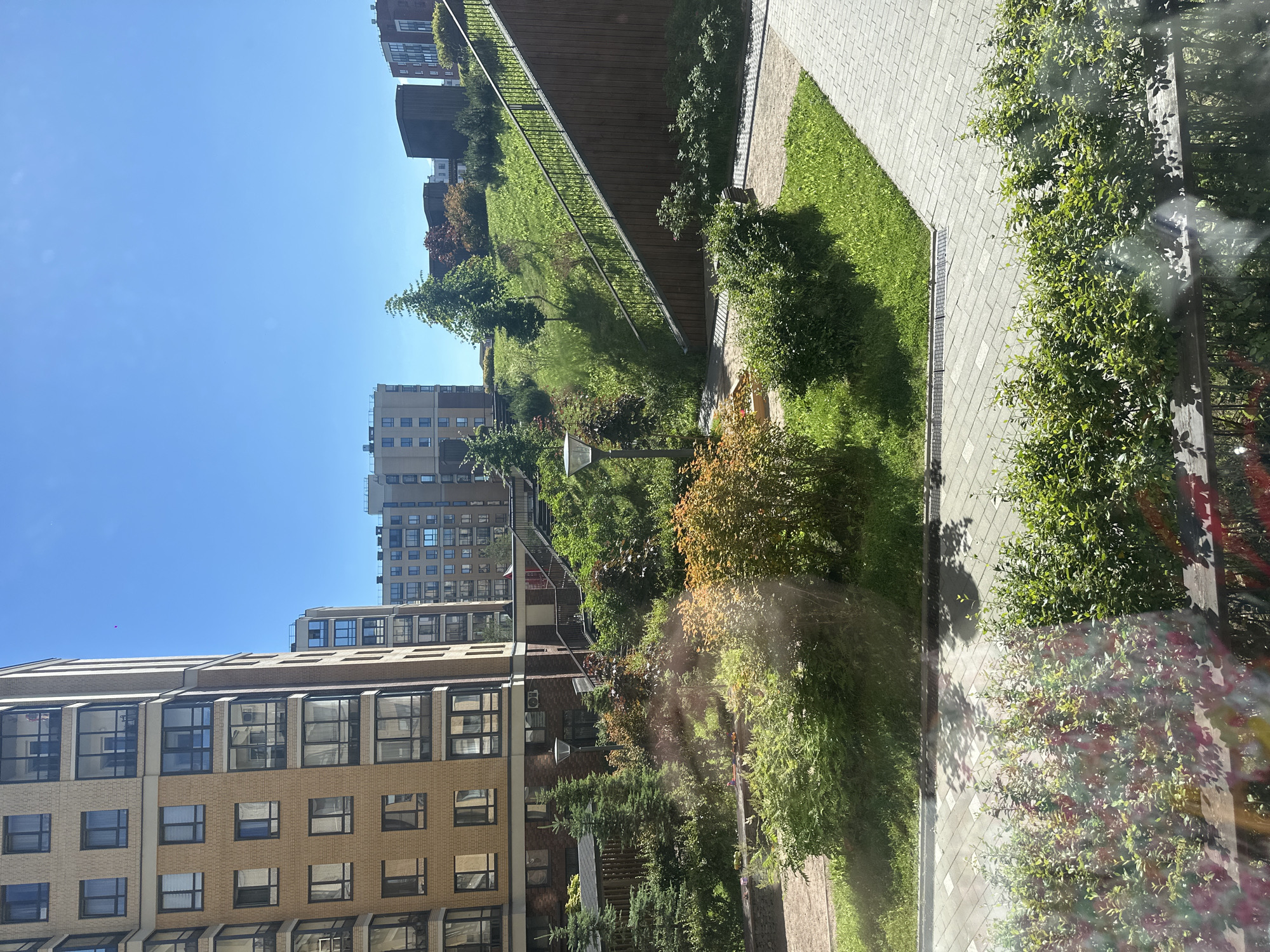 This media file depicts an outdoor scene featuring modern apartment buildings surrounded by landscaping. The buildings are multi-story structures with many windows, constructed with materials like brick and glass. In the foreground and mid-ground, there are areas of green grass, paved walkways, and various types of trees and bushes, some with autumnal colors. A sloped grassy area is visible, bordered by a wooden fence or wall and a metal railing. The sky is clear and blue, indicating daytime and fair weather. There are no people or animals visible in the frame. The scene appears to be a well-maintained residential complex or urban development. There is no discernible text in the image.