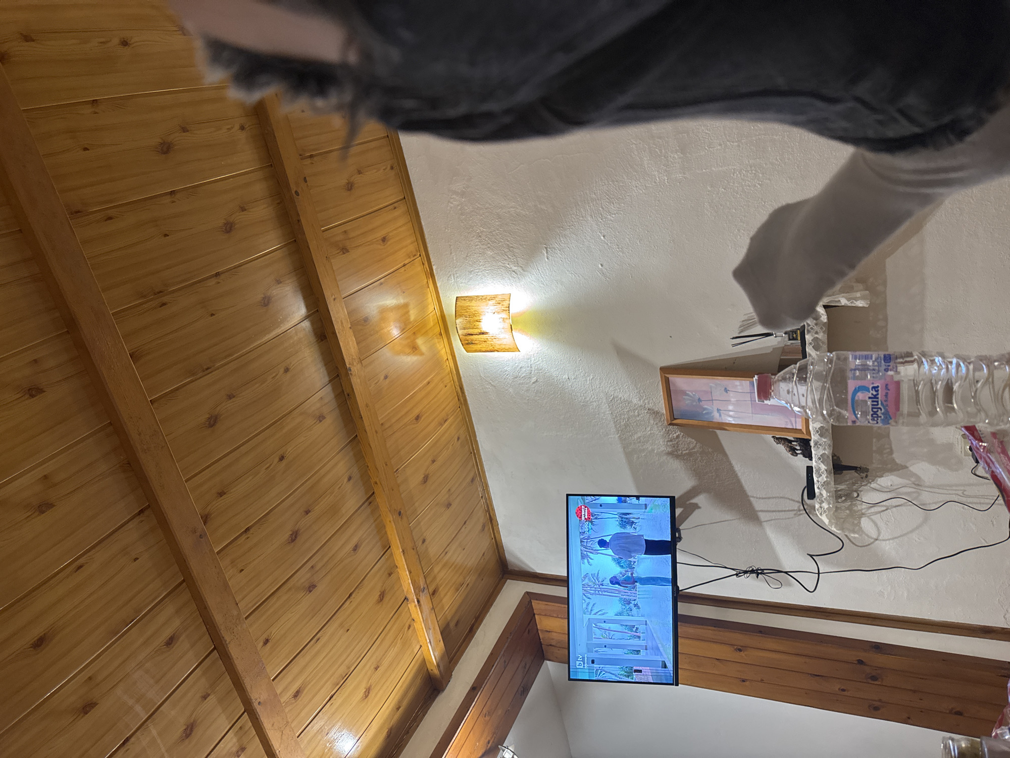 The image shows an interior space with a pitched wooden ceiling, a white wall with a wall sconce, and a television mounted on the wall displaying a scene with people on a beach. There is a framed picture above the television, and a water bottle is placed on a decorative shelf. The foreground contains a blurred view of clothing or bedding. The location is Yambol, Bulgaria.