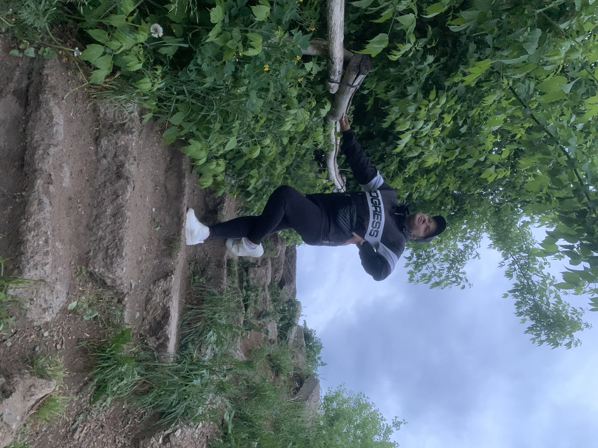 A person in dark athletic wear and white sneakers is standing on a dirt path with steps leading up a small, grassy incline. The person is holding onto a branch extending from foliage, and the sky is visible through the leaves of the trees. The location is Chaikovsky, Russia.