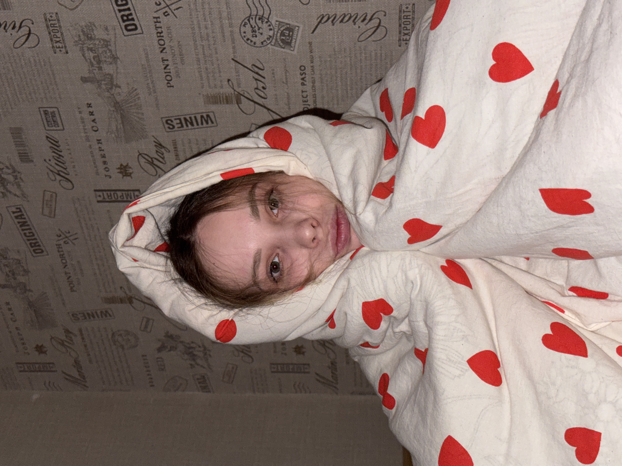 A young woman with light skin and brown hair is wrapped in a white blanket patterned with red hearts. She is lying down, and her face is visible, looking towards the camera. The background consists of a textured wallpaper or fabric with a repeating pattern of wine-related labels and graphics. The location is indeterminable from the visible information, and thus cannot be said to be Chaikovsky, Russia. There are no discernible actions or interactions.