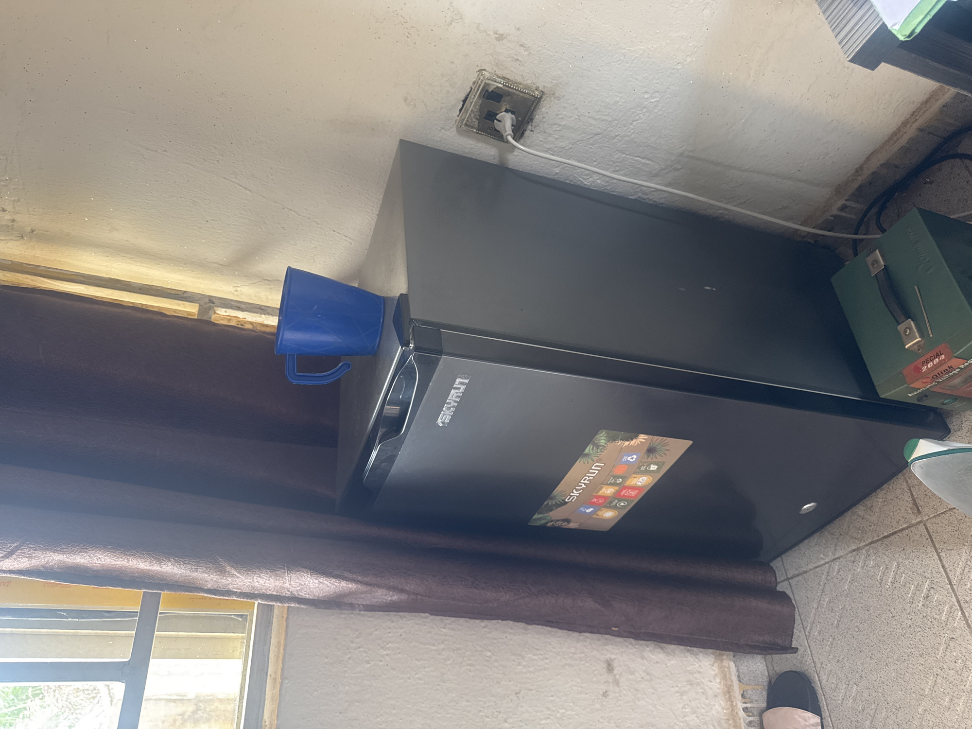 An indoor shot depicts a small black refrigerator, identified by the "SKYRUN" logo, positioned in a corner near a window with a brown curtain. A blue plastic cup rests on top of the refrigerator. A white power cord extends from the refrigerator to a wall outlet, which appears to be damaged and exposed. The walls are textured and off-white, with some visible dust or debris. The floor is tiled with a light gray pattern. A pair of black and pink slippers are visible on the left side of the frame. A green box with a handle and some labels is partially visible on the floor in the lower right quadrant, and a stack of papers is on a surface to the right. The lighting suggests it is daytime, with light filtering through the window.