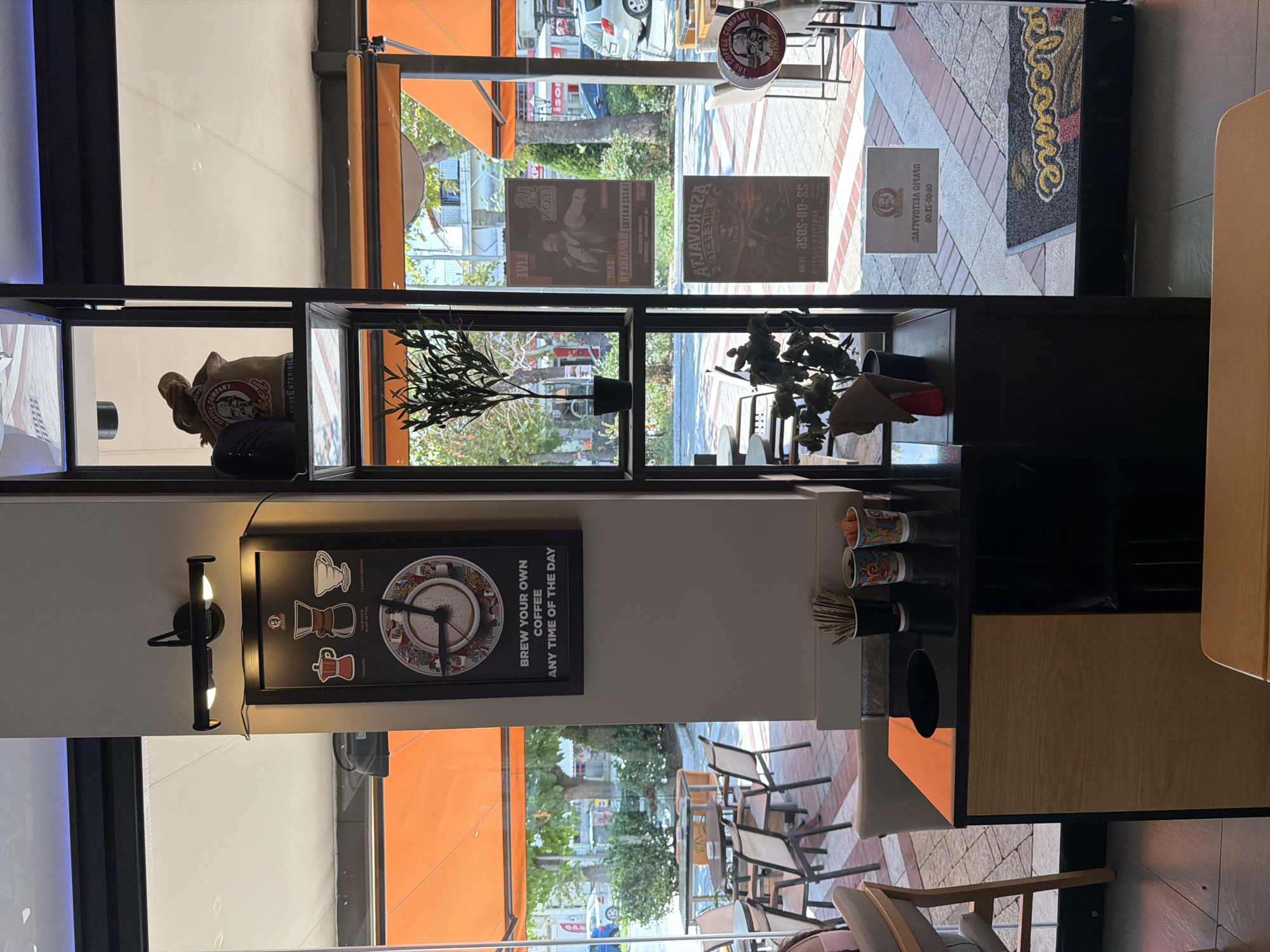 The image depicts the interior of a cafe in Asprovalta, Greece, with a clear view of the exterior through a large window. The cafe features a modern, minimalist design with dark metal shelving units. On one shelf, a burlap sack with "COFFEE COMPANY" and a portrait of a man is displayed. Below this, a framed black clock with coffee-related imagery and the text "BREW YOUR OWN COFFEE ANY TIME OF THE DAY" is hung on a light-colored wall. Another shelf holds a small potted olive tree.
Adjacent to the shelving unit is a dark countertop with various items, including cups containing straws and decorative items. A wooden cabinet with a cutout for waste disposal is positioned beneath the countertop. Through the window, outdoor seating areas with tables and chairs are visible, along with the street and buildings of Asprovalta. Several posters are affixed to the window. One poster, visible from the inside, features a man in vintage attire. Another, also seen from the inside, displays a motorcycle. A circular sign for "THE COFFEE COMPANY" with a man's portrait is attached to a pillar. A welcome mat with "welcome" written on it is visible on the floor near the entrance. The overall atmosphere suggests a well-lit and inviting cafe environment.