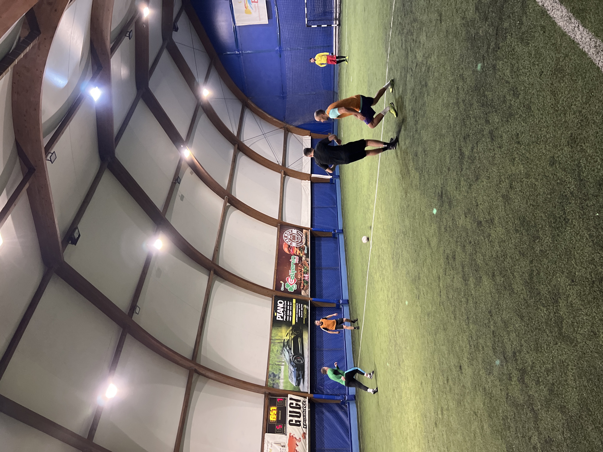 The image depicts an indoor soccer field, possibly in Budva, Montenegro, due to the presence of Cyrillic text on advertising boards. The building has a high, arched roof supported by wooden beams and is illuminated by overhead lights. Several individuals are on the field: two men in black and orange gear practice drills near the center. One man in yellow stands near a goal in the distance. A soccer ball sits on the field. One man in green and one in orange are near the advertising boards.