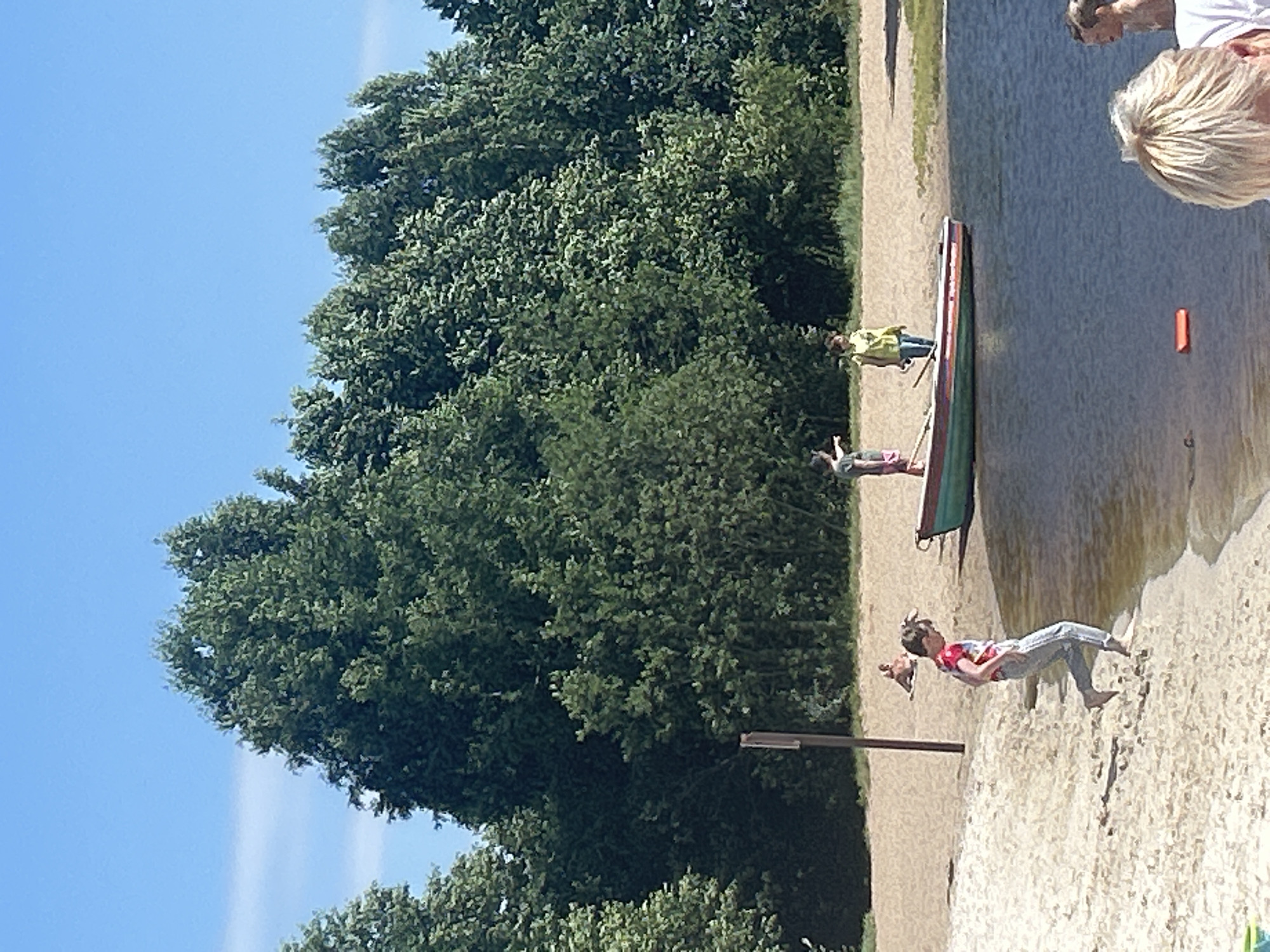 A sunny day at the beach in Sestroretsk, Russia, with people enjoying the water and sand. In the foreground, a young child in a red and white t-shirt and jeans is running along the water's edge on the sand. Further back, a green and red rowboat is docked by the shore. Two individuals are standing near the boat, one pointing towards the trees. In the background, lush green trees line the beach. Several other people are visible lounging on the sand in the distance. The water is calm and reflects the bright blue sky.