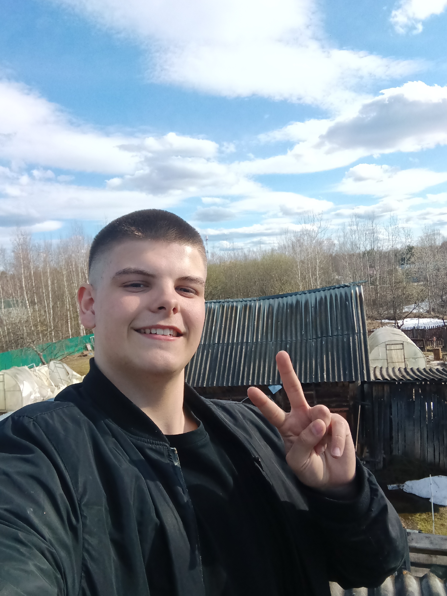 A young man takes a selfie outdoors on a partly cloudy day. He is wearing a black jacket and a black shirt. He is smiling and holding up two fingers in a peace sign. Behind him are wooden buildings, a greenhouse, and trees. There is some snow on the ground. The location is most likely in or around Красный Маяк, Russia.